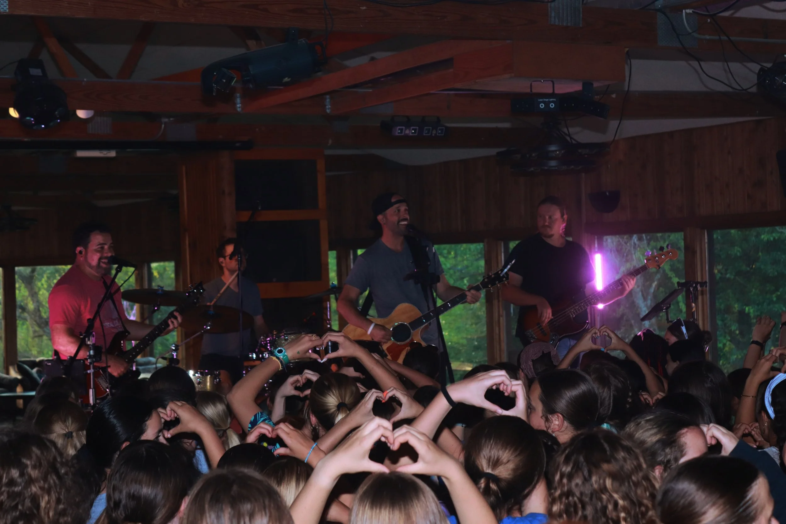 A band performing on stage at an indoor venue with a crowd of people in front making heart shapes with their hands.