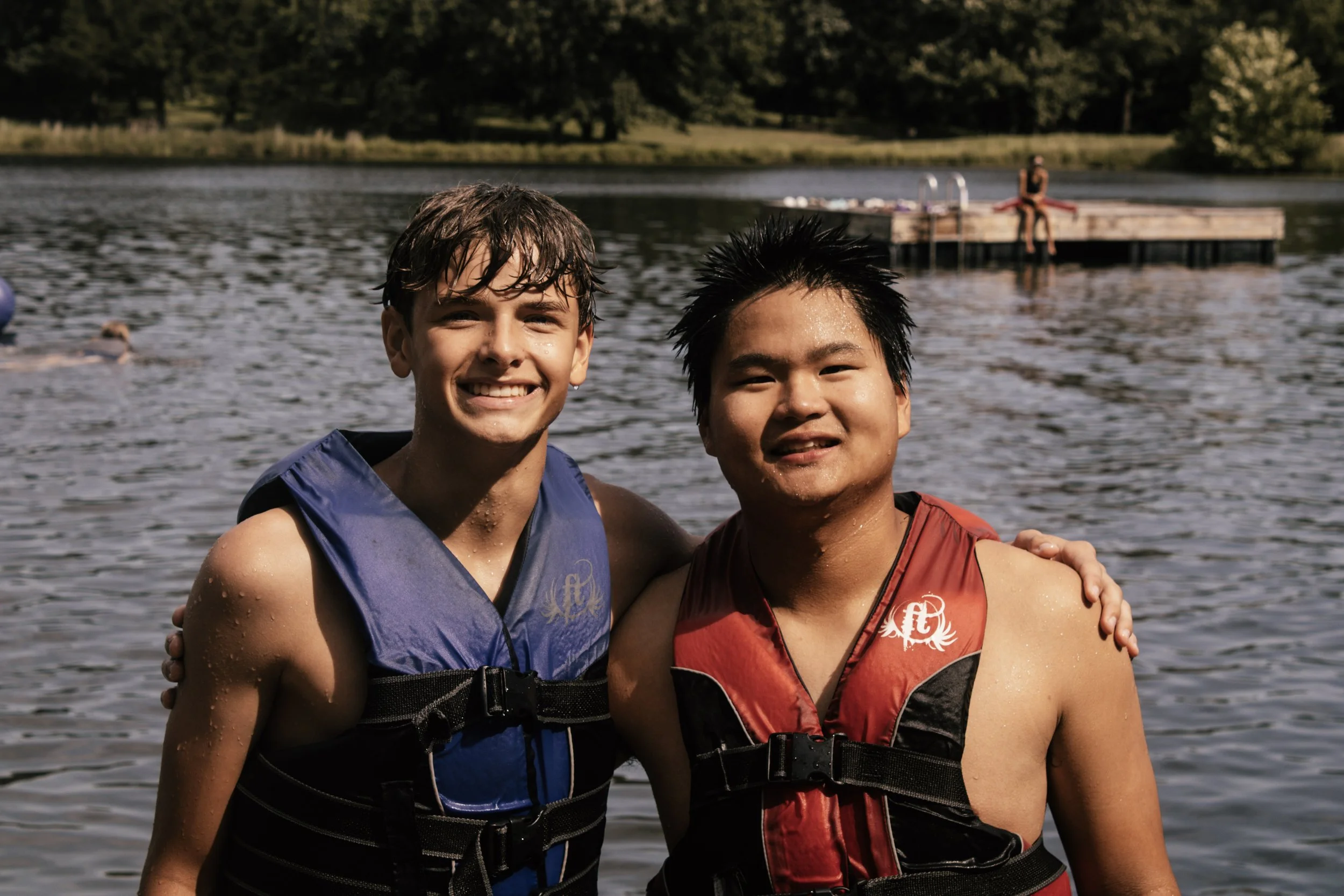 Two boys smiling and standing close together near a lake, wearing life jackets, with a dock and a person in the background