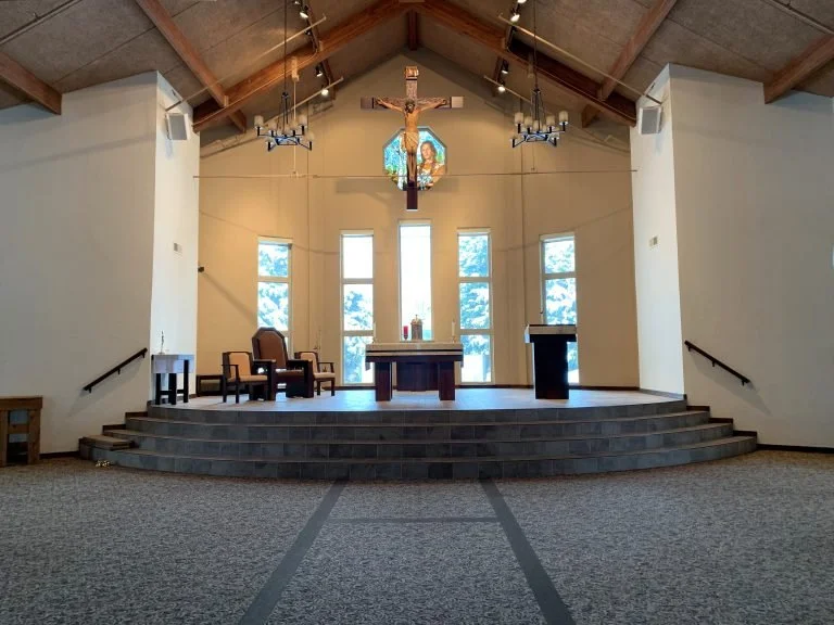 Interior of a church with an altar, crucifix hanging above, stained glass window depicting Jesus, and warm lighting from chandeliers.
