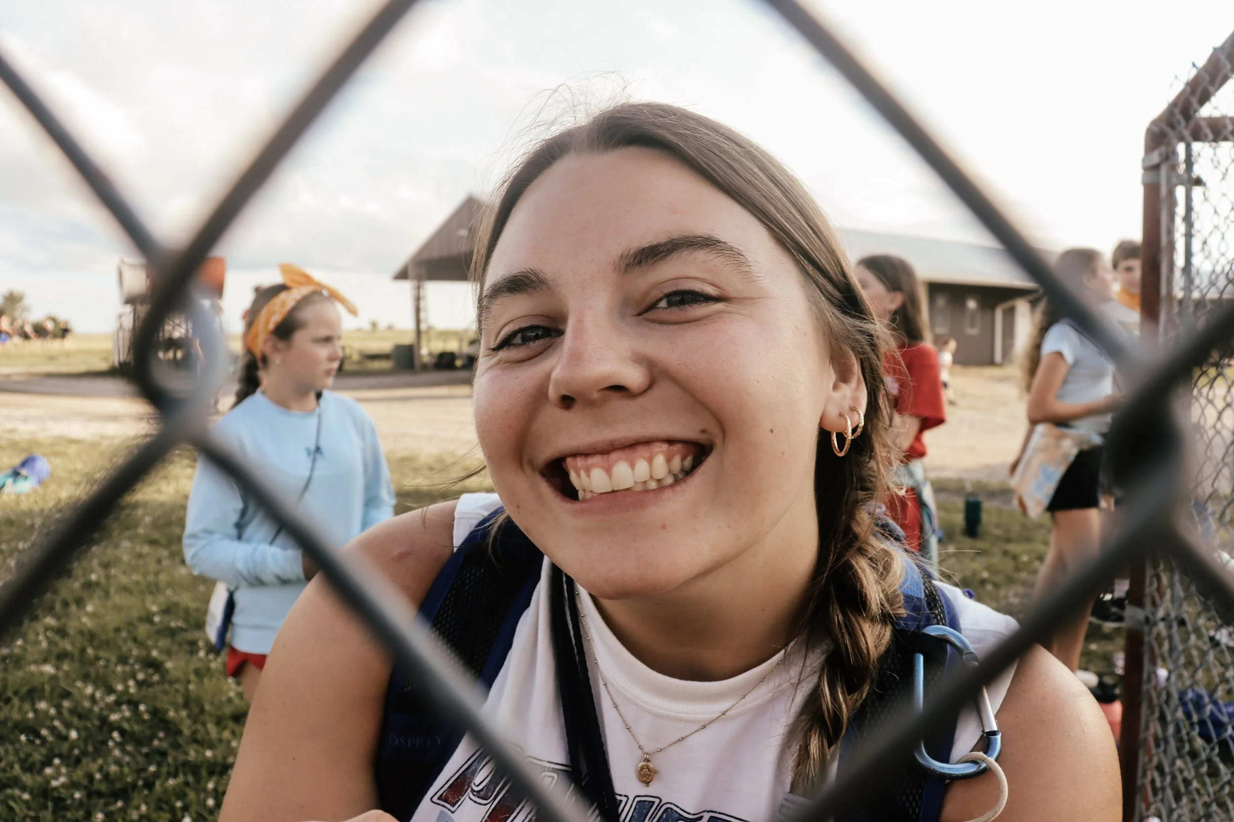 Close-up of a smiling young woman with light brown hair, hoops earrings, and wearing a white shirt and backpack, seen through a chain-link fence with a grassy field and other children in the background.