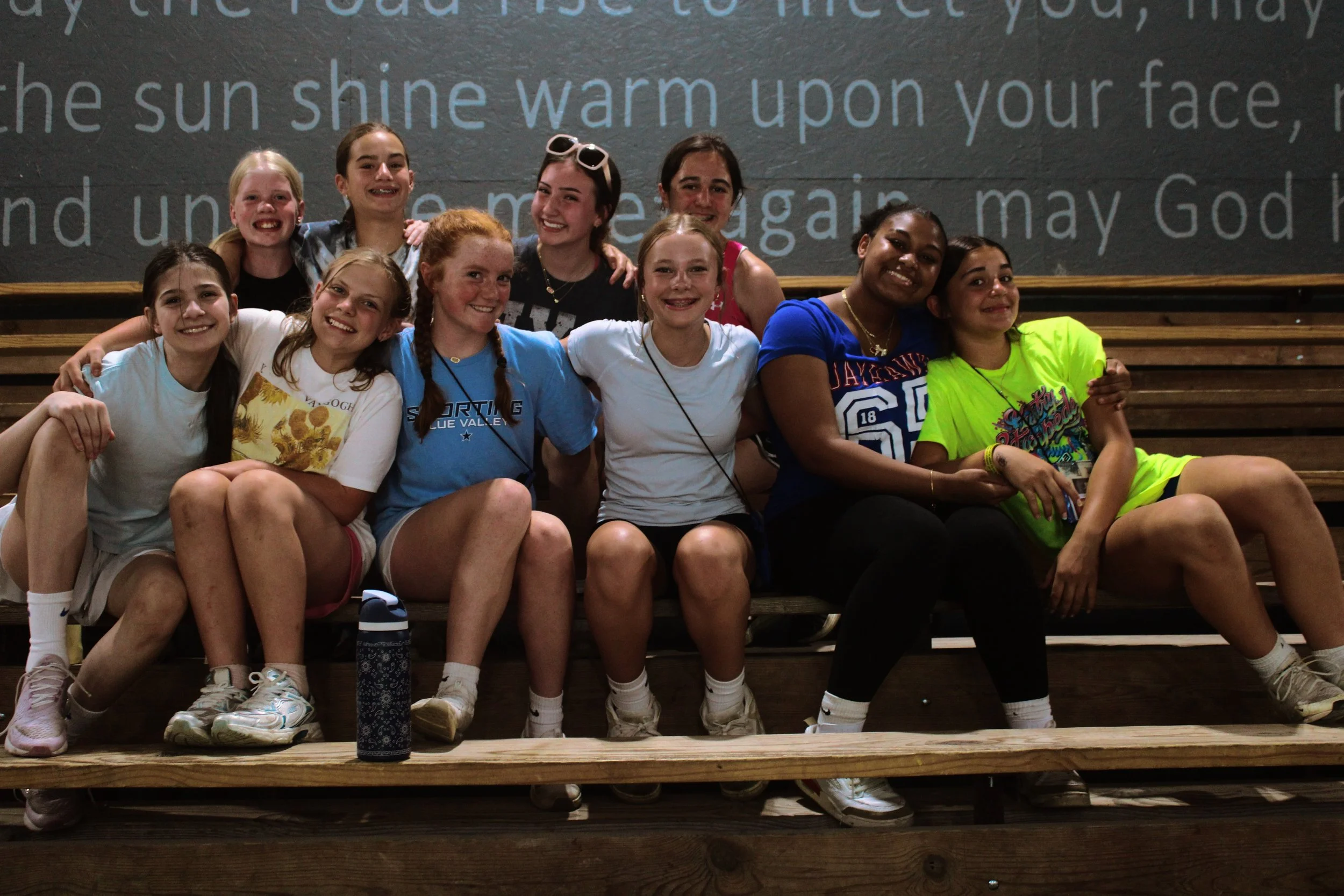 Group of children sitting on wooden bleachers indoors, smiling and posing for the camera, with text projected on the wall behind them.