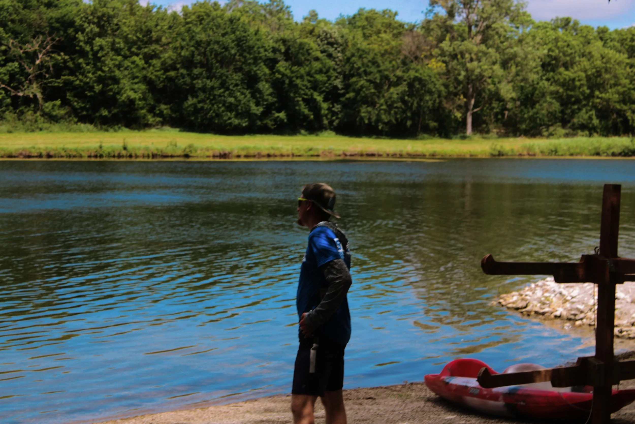 Person in blue jacket and black shorts standing by a lake, with trees in the background and a red paddle boat nearby.