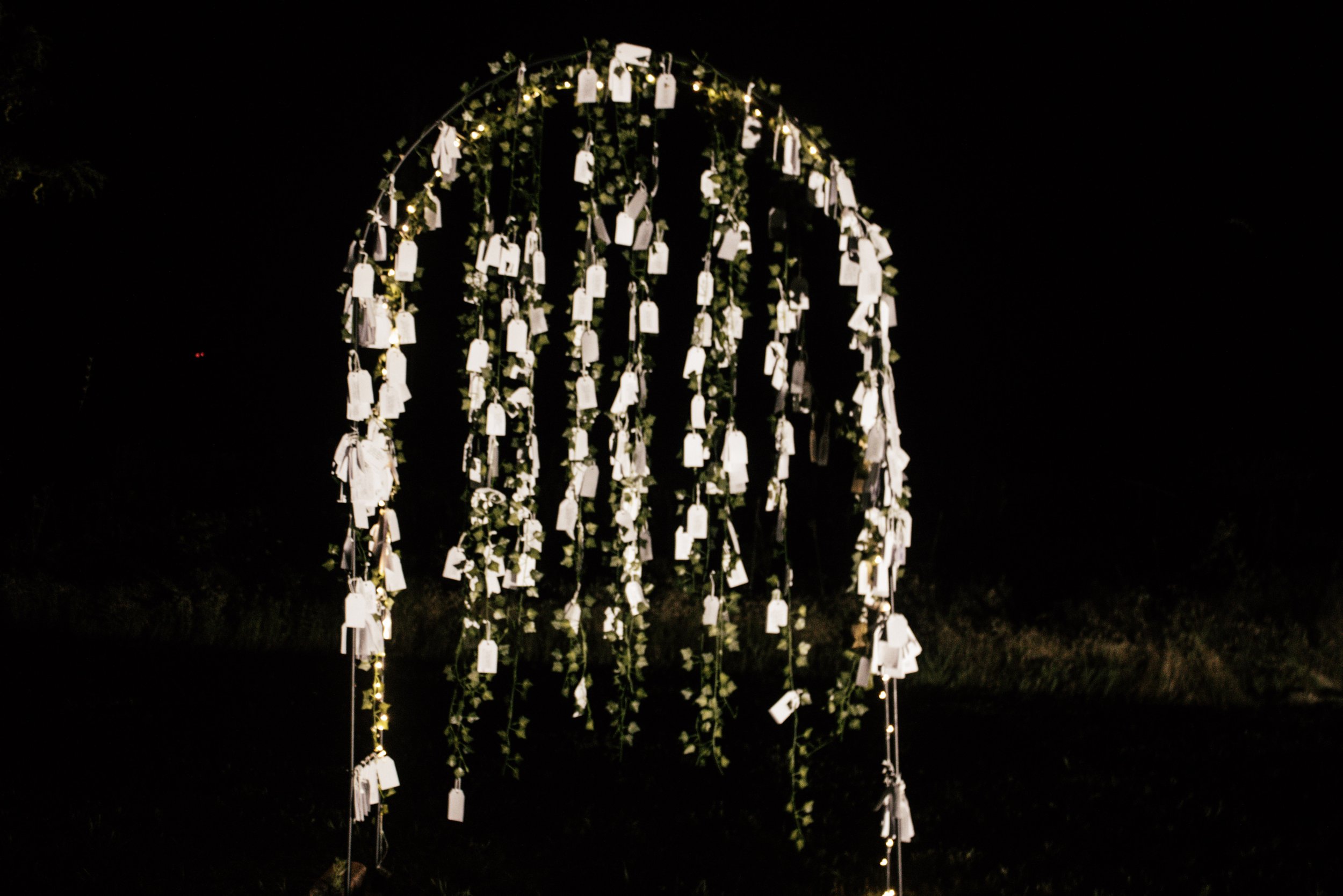 Nighttime outdoor scene featuring a decorative metal arch with hanging white tags or lanterns, adorned with small lights and green foliage.