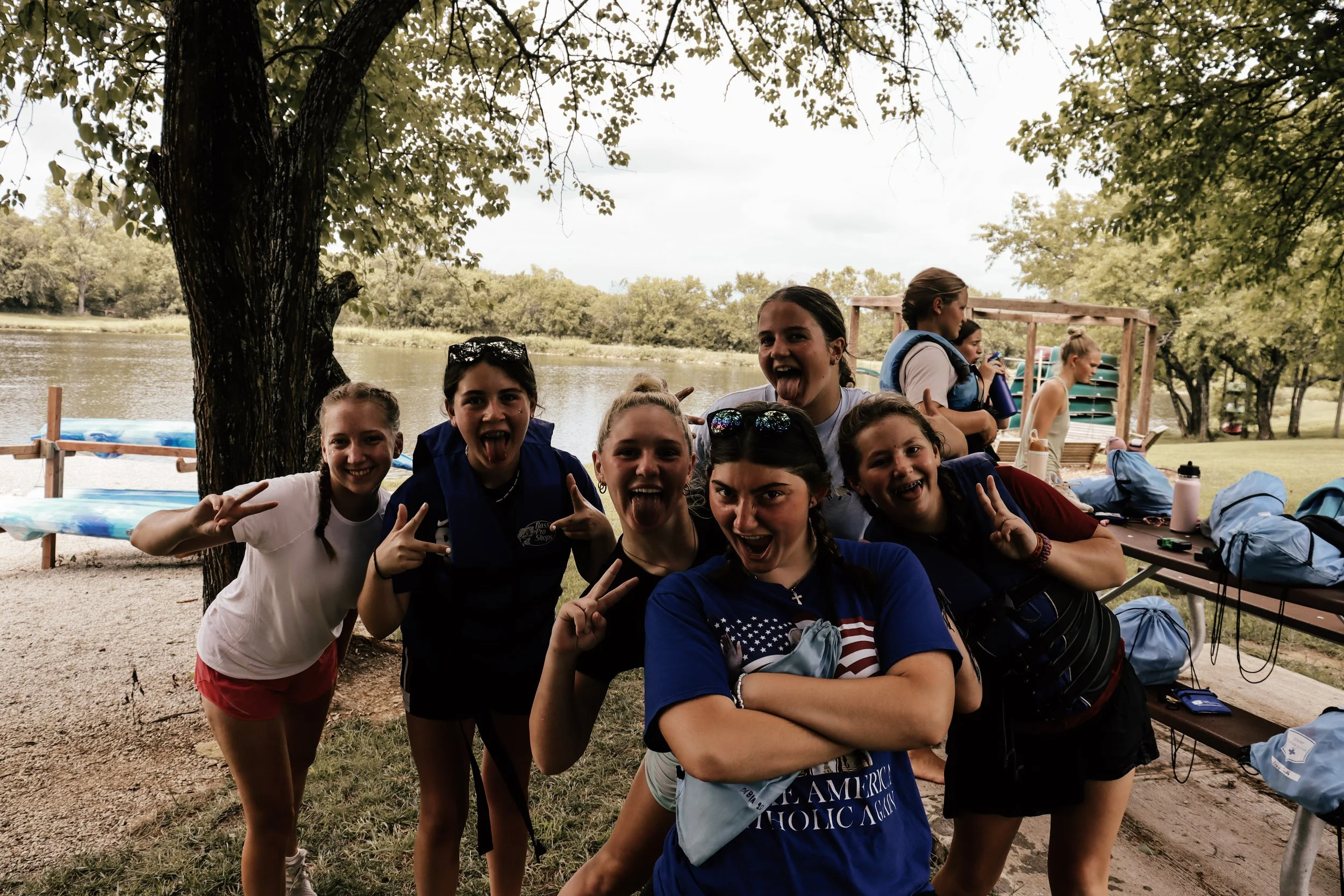 Group of smiling young girls making peace signs and having fun outdoors near a lake with trees and a picnic area in the background.