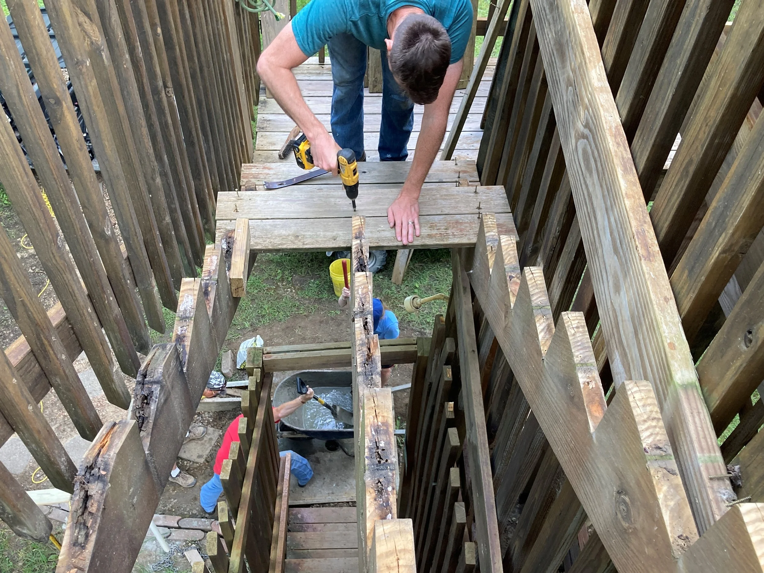 People working on building a wooden deck, with one person using a drill and others working underneath through an opening in the deck.