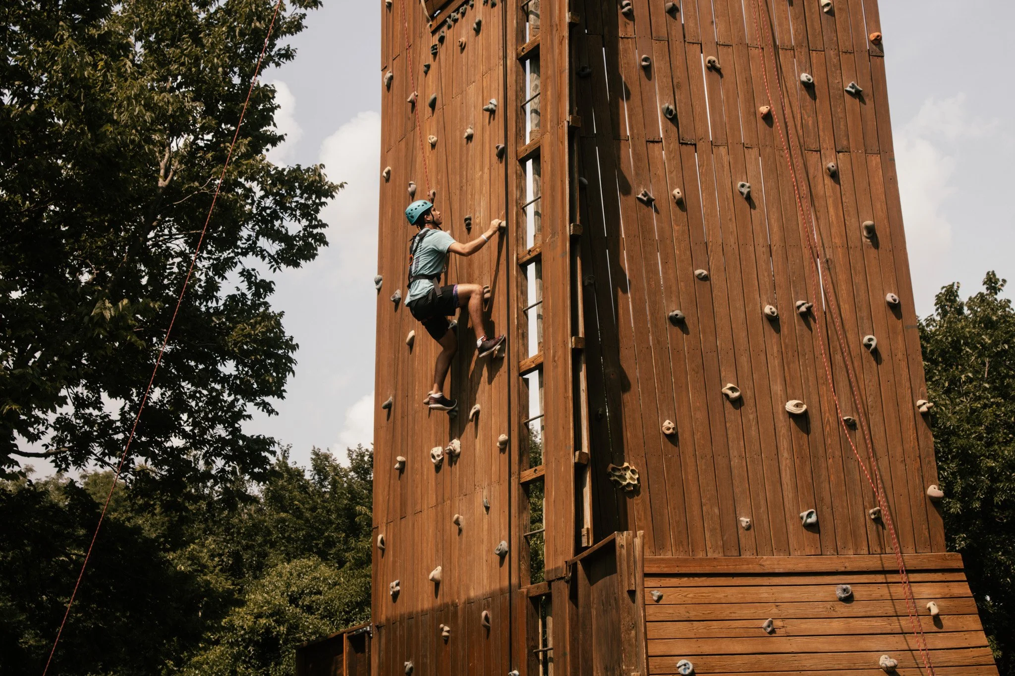 A person wearing a helmet and harness climbing a wooden outdoor rock climbing wall.