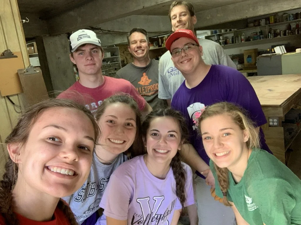 A group of eight young people and one adult in a workshop or garage, smiling for a group selfie. Shelves with tools and supplies are visible in the background.