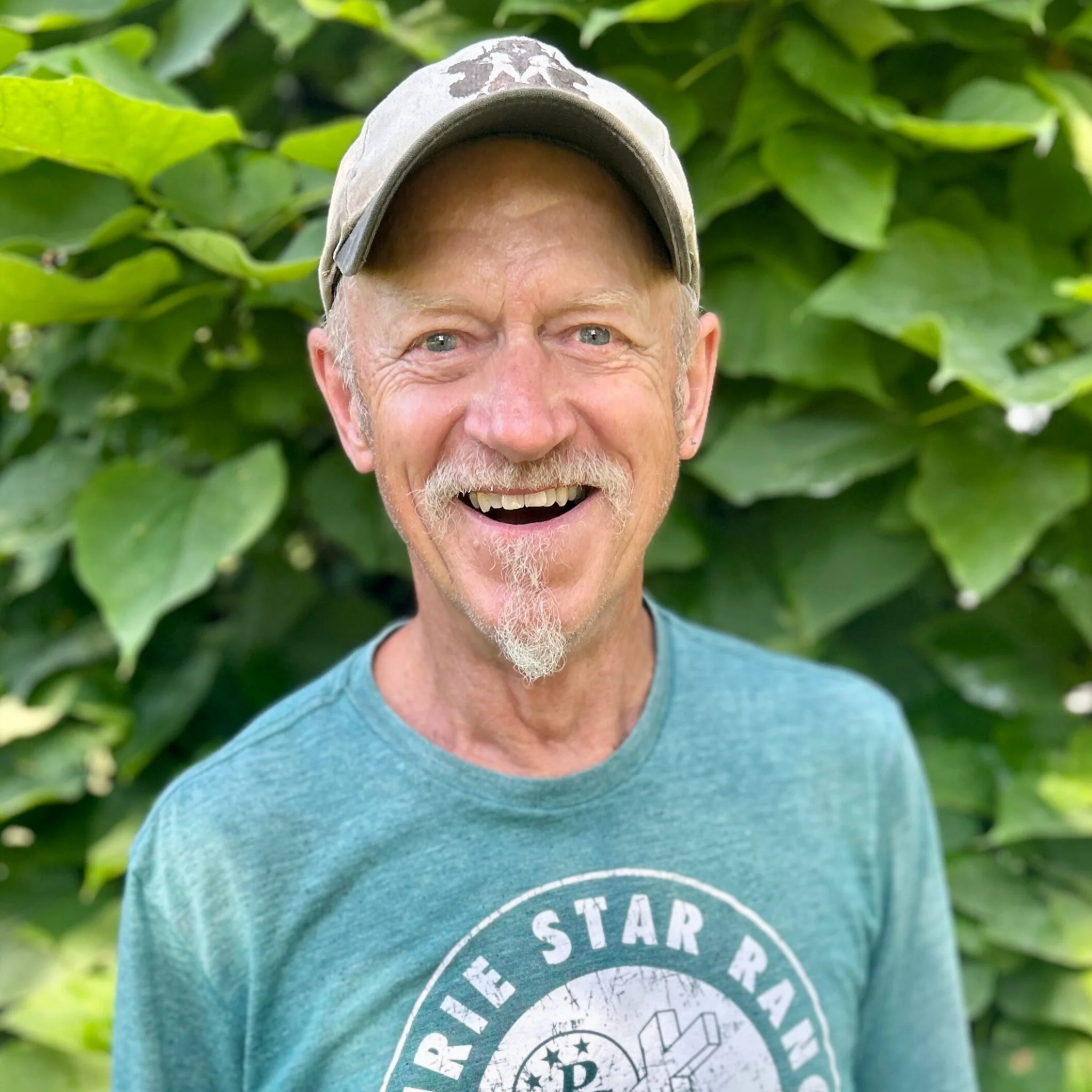 Close-up of a smiling man with a beard wearing a gray baseball cap and a teal t-shirt, standing in front of green foliage.