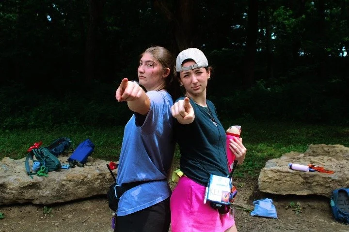 Two teenage girls standing back-to-back outdoors near a wooded area, pointing at the camera. One girl is wearing a blue shirt and a white cap, holding a pink water bottle, while the other is in a black shirt and pink shorts, with a cap backwards. Bac