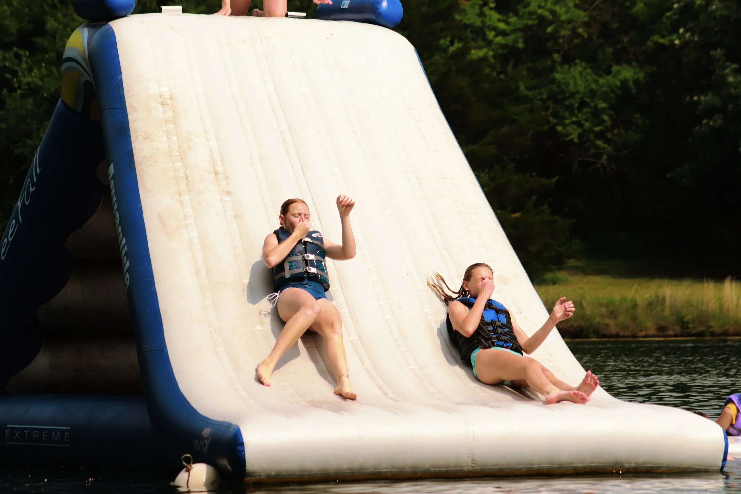 Two girls in life jackets sliding down an inflatable water slide into a lake, surrounded by trees.