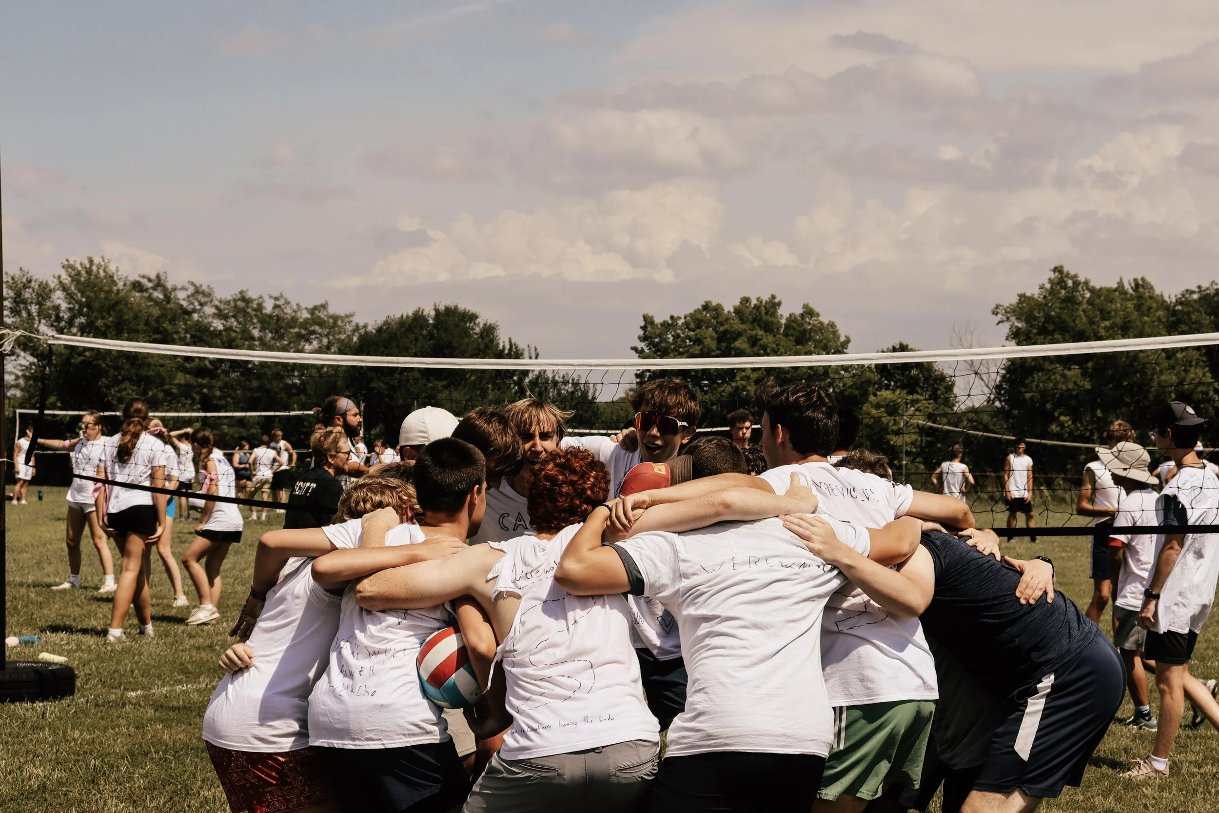 A group of people in white T-shirts with writing on them huddle together in a circle outdoors during daytime, celebrating after a volleyball game on a grassy field with trees and a partly cloudy sky in the background.