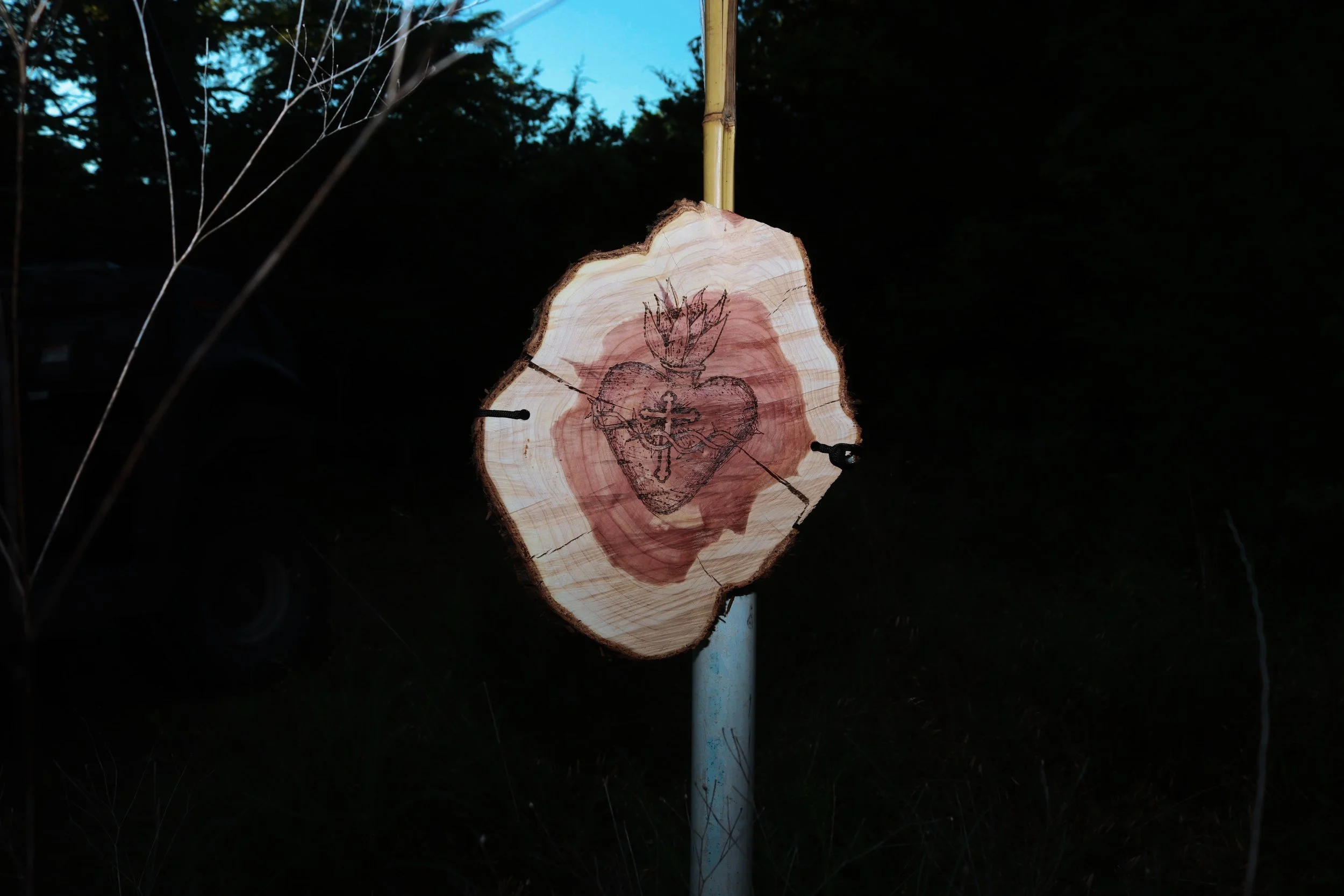 A circular wooden plaque with a drawing of a heart pierced by a sword and a fire burning above it, mounted on a white pole outdoors at dusk or night.