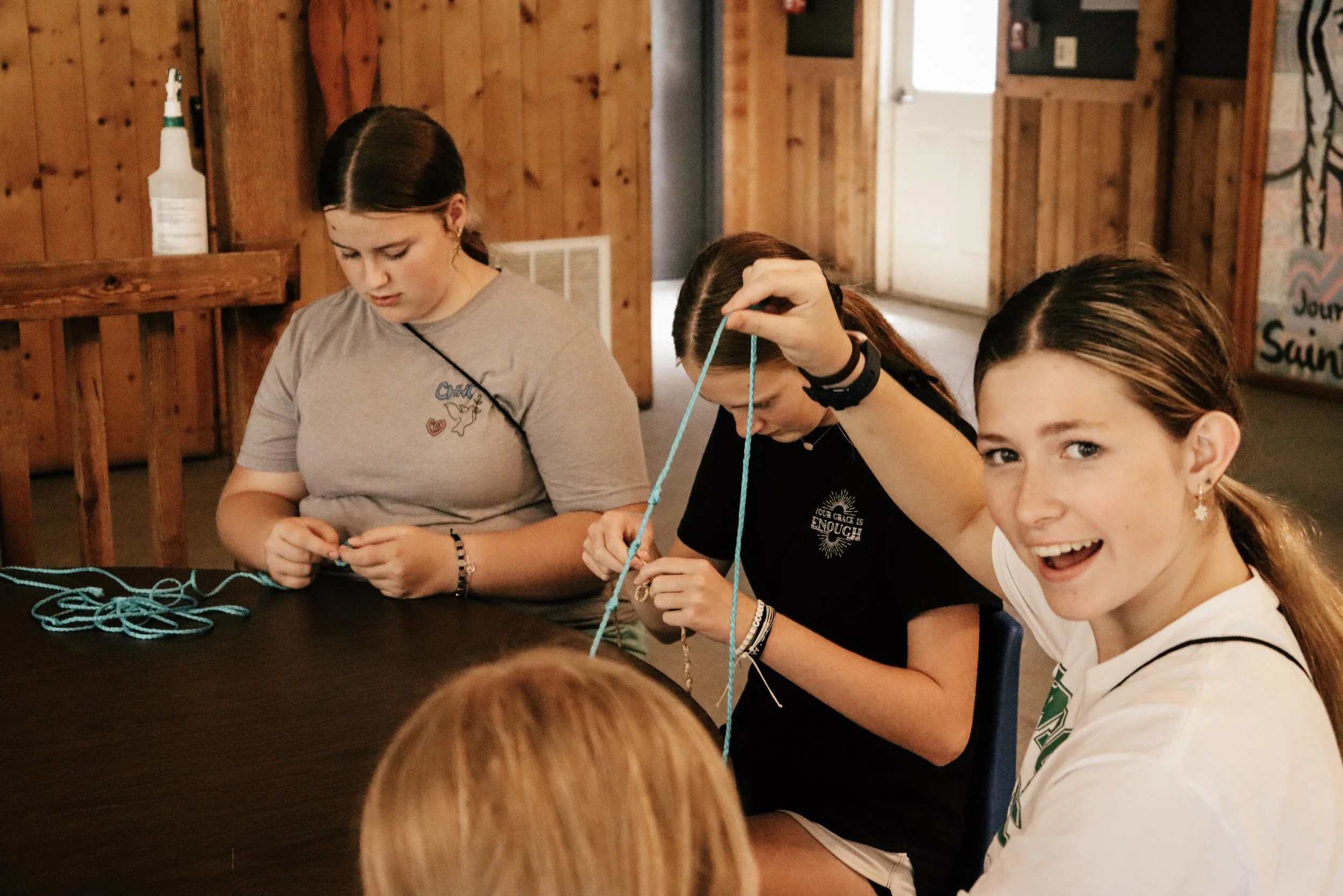 A group of girls sit around a table, working on a craft project with blue string, in a wooden interior room. One girl is smiling at the camera, revealing braces, while the others focus on their work.