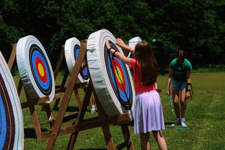 A girl in a red shirt and purple skirt throws an axe at a large indoor archery target outside during daytime. Other people are in the background on a grassy field.