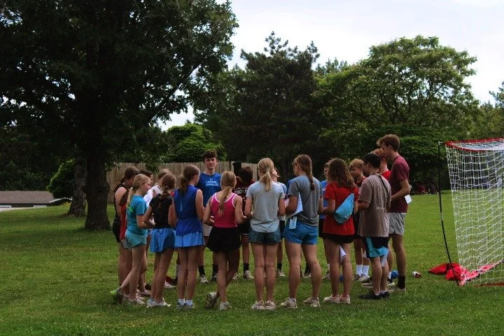 A group of children and teenagers gathered in a circle on a grassy field during daytime, with a soccer goal and trees in the background.