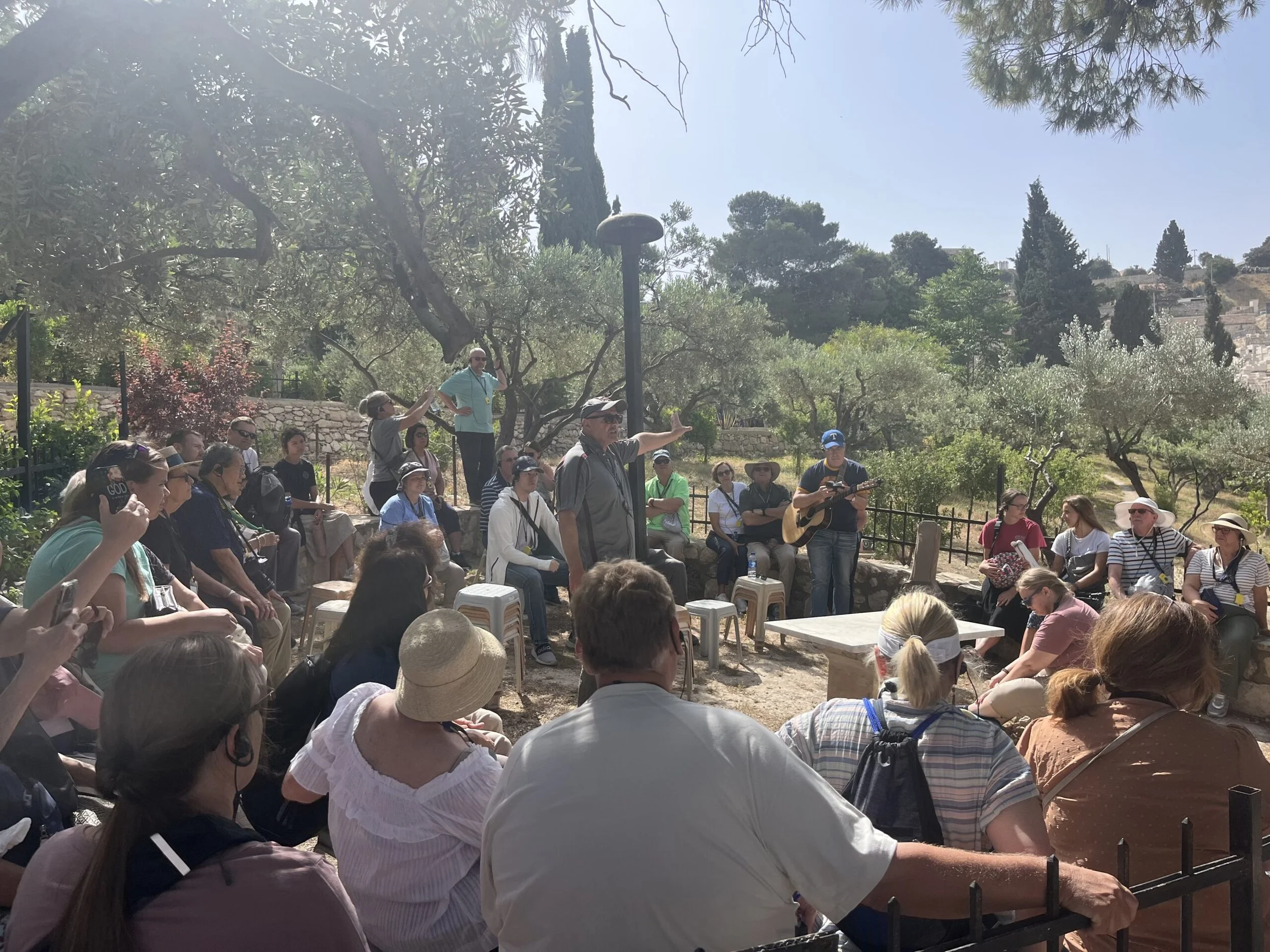 A group of people gathered outdoors, listening to a man speaking and playing guitar, surrounded by trees on a sunny day.