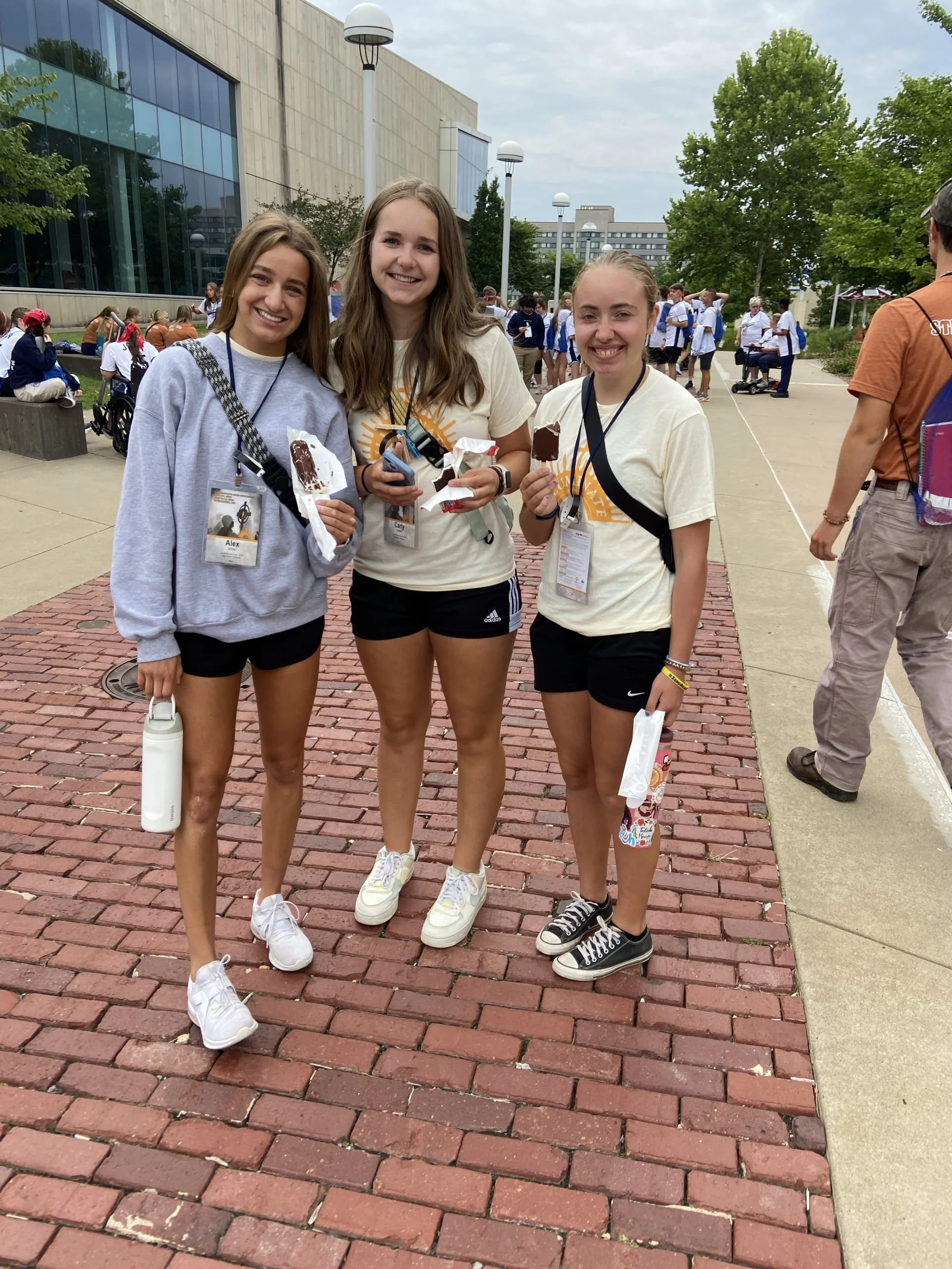 Three young women standing outdoors on a sidewalk, smiling at the camera. They are wearing casual athletic clothing and have name tags around their necks. In the background, there are more people sitting and standing, some wearing similar attire, nea