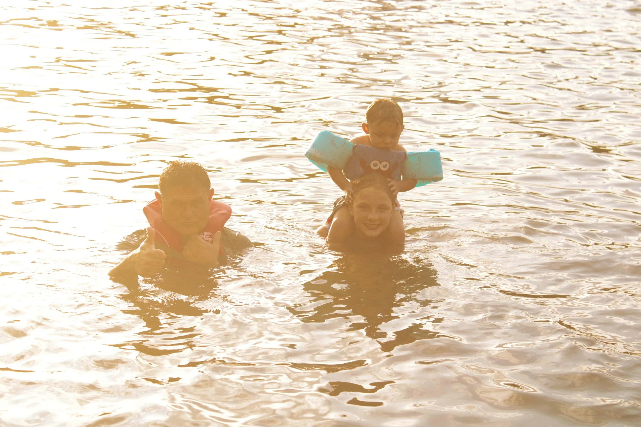 Three people in a lake or river during sunset, including a smiling woman giving a thumbs-up, a young boy with water wings on his arms sitting on the woman's shoulders, and a man in the water giving a thumbs-up.
