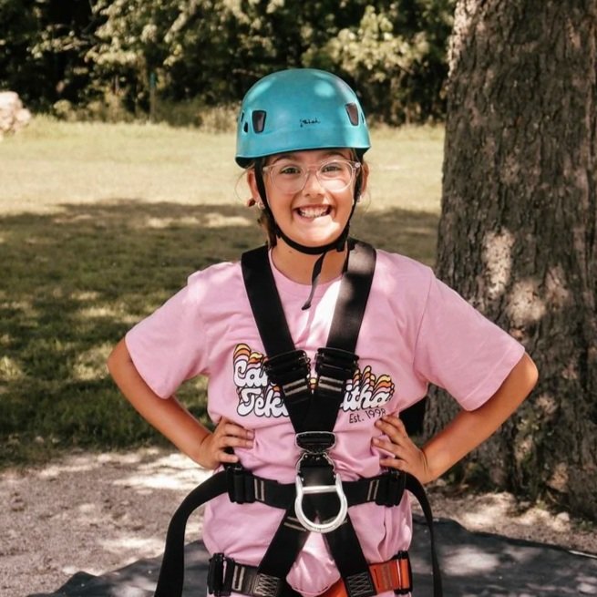 A young girl wearing a blue helmet, pink t-shirt, and safety harness outdoors near a tree on a sunny day.