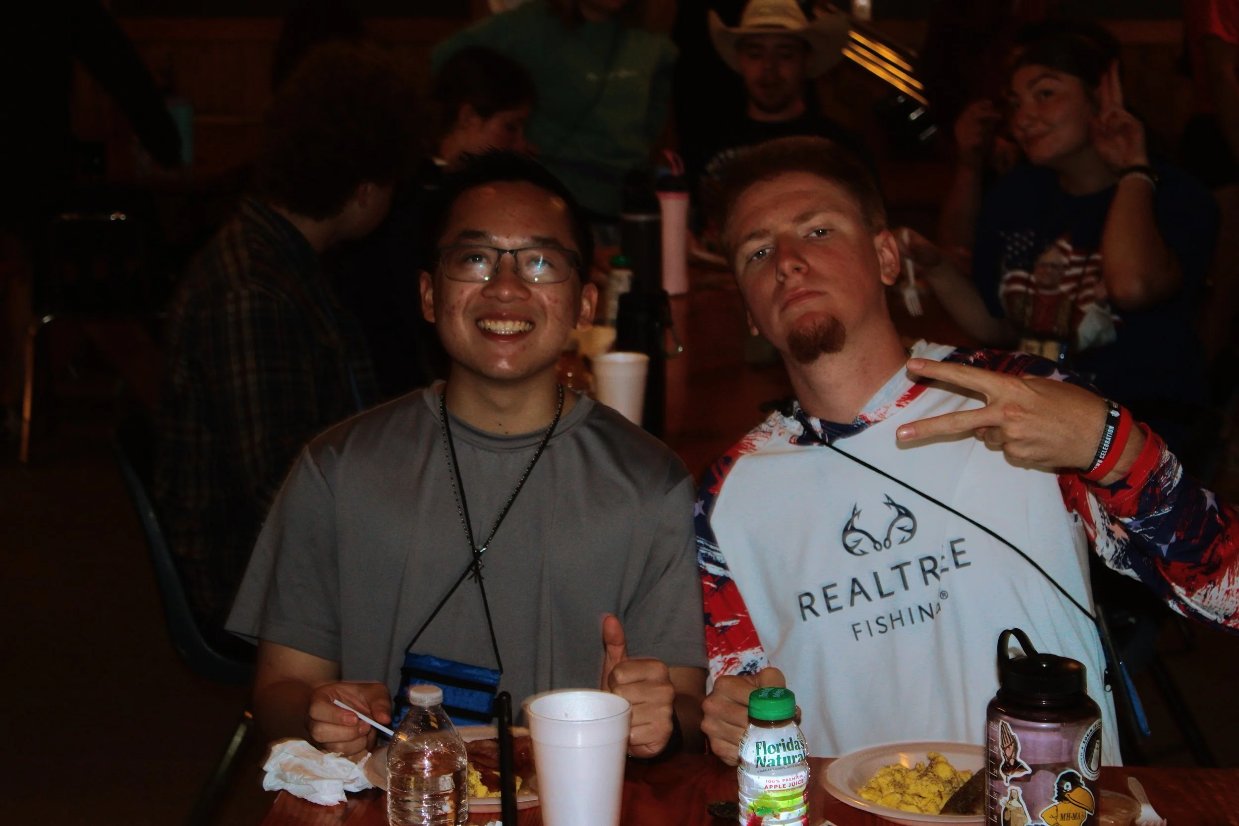 Two young men sitting together at a table in a dark indoor setting, smiling at the camera. The man on the left is wearing glasses and a gray T-shirt, while the man on the right is making a peace sign and wearing a white, red, and blue patriotic shirt