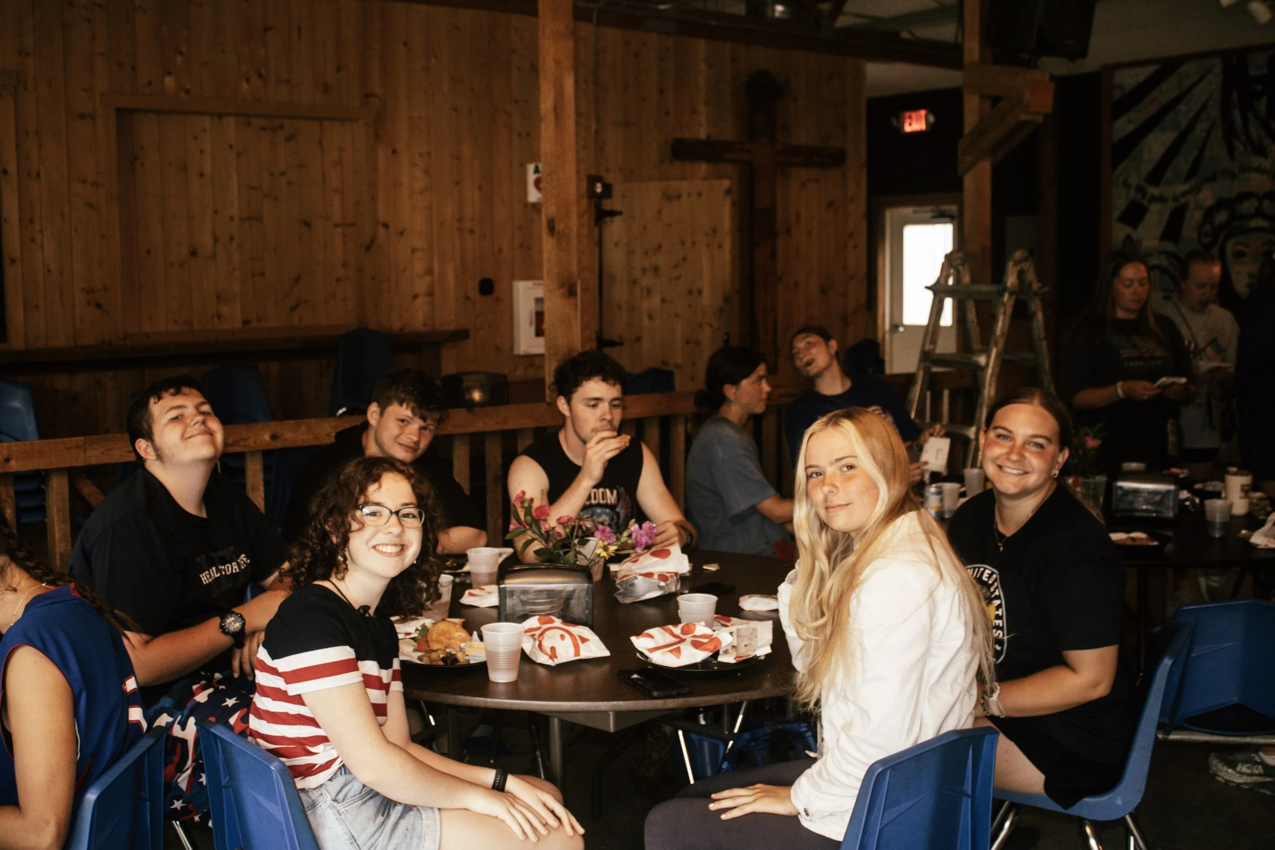 Group of young people sitting at a table during a meal at indoor event with wooden walls and mural artwork.