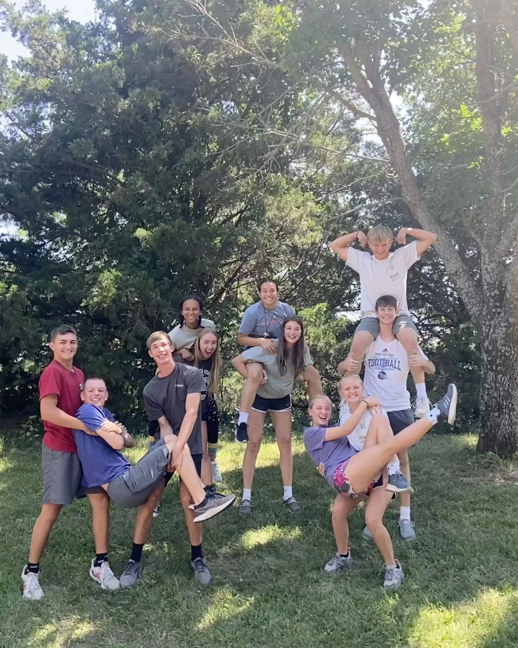 Group of teenagers outdoors forming a human pyramid and smiling, with trees and grass in the background.