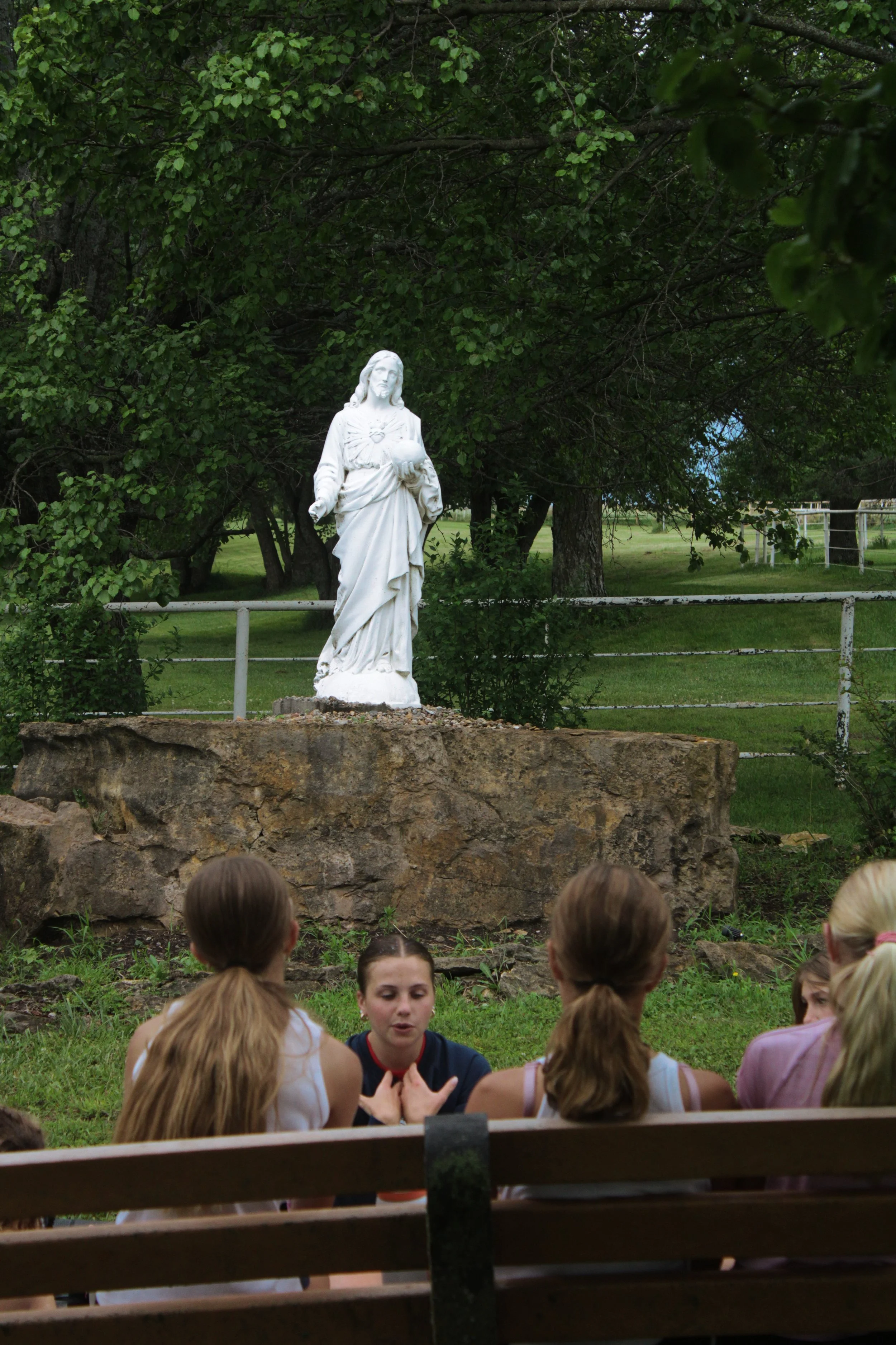 A white statue of Jesus Christ holding a heart, situated outdoors in front of green trees and grass, with children and an adult seated in front of it on a bench.