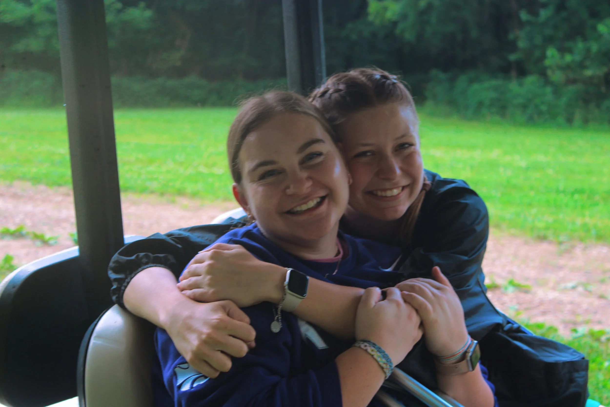 Two girls hugging and smiling inside a vehicle with a green outdoor background.