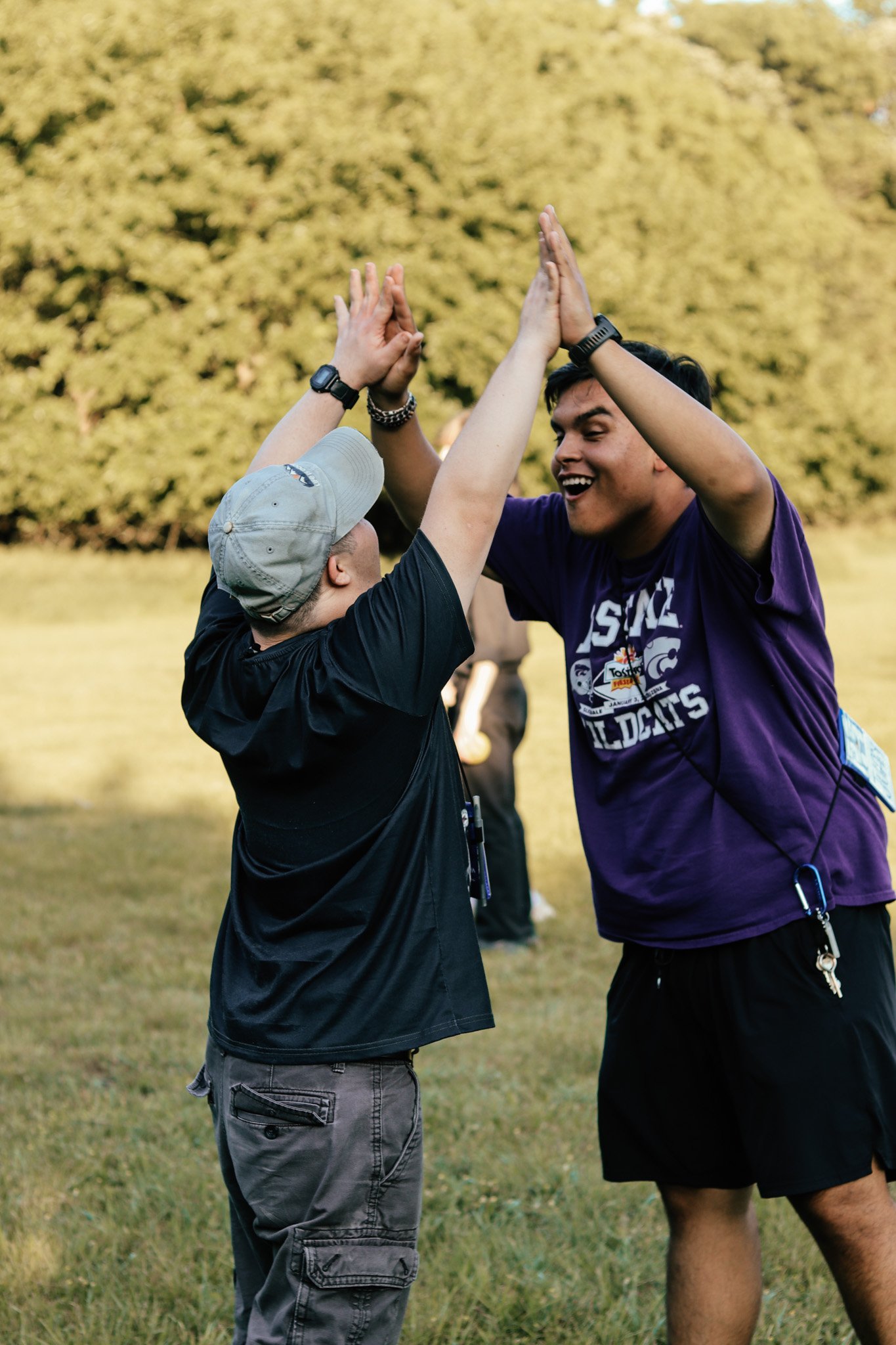 Two young men high five outdoors on grass, with trees in the background. One is wearing a baseball cap, black shirt, and gray shorts, while the other is wearing a purple T-shirt and black shorts. They are smiling and appear happy.