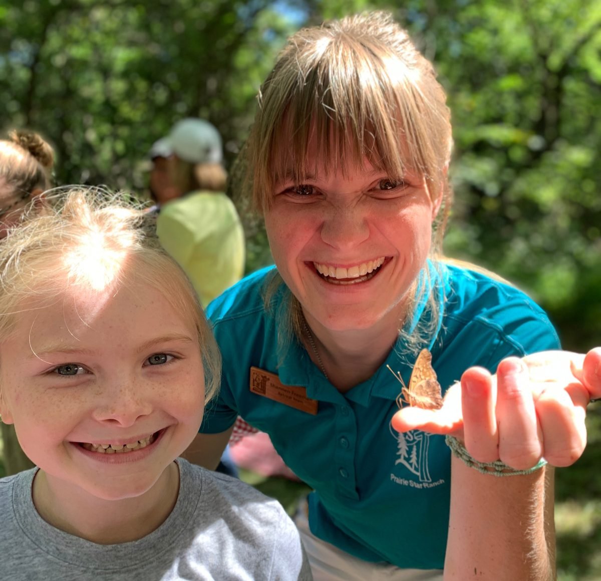 A smiling woman holding a butterfly on her hand next to a young girl with blonde hair and freckles, both outdoors in a green forested area.