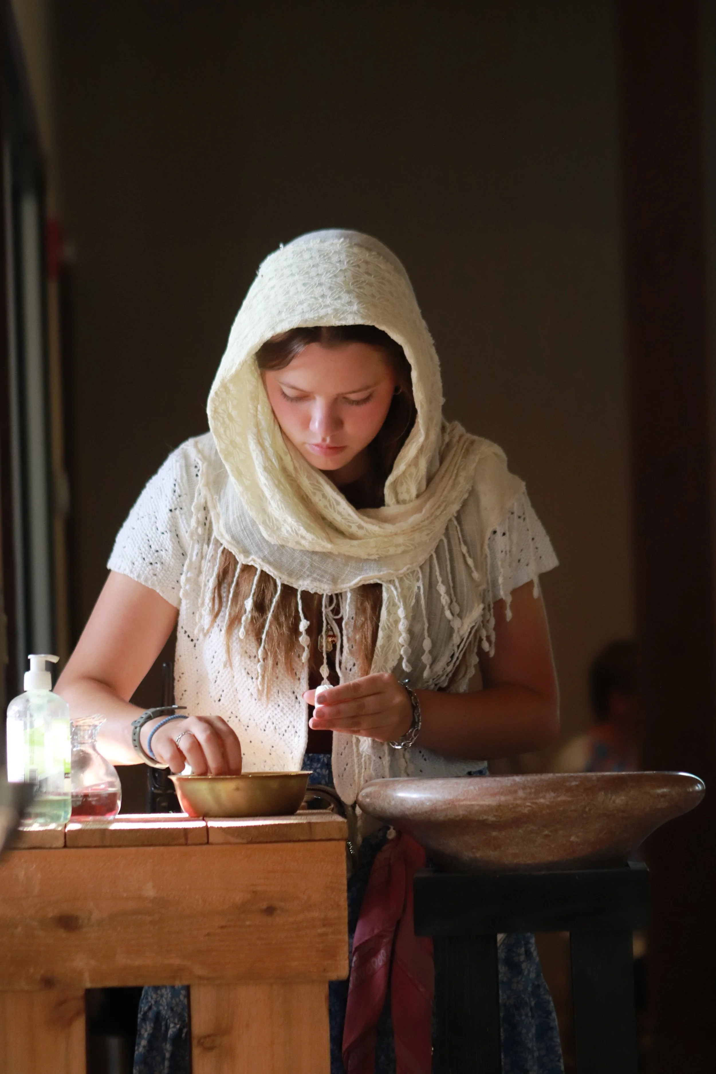 A young woman wearing a cream lace hood over her head, focused on a task at a wooden counter with various bottles and containers, in a dimly lit room.