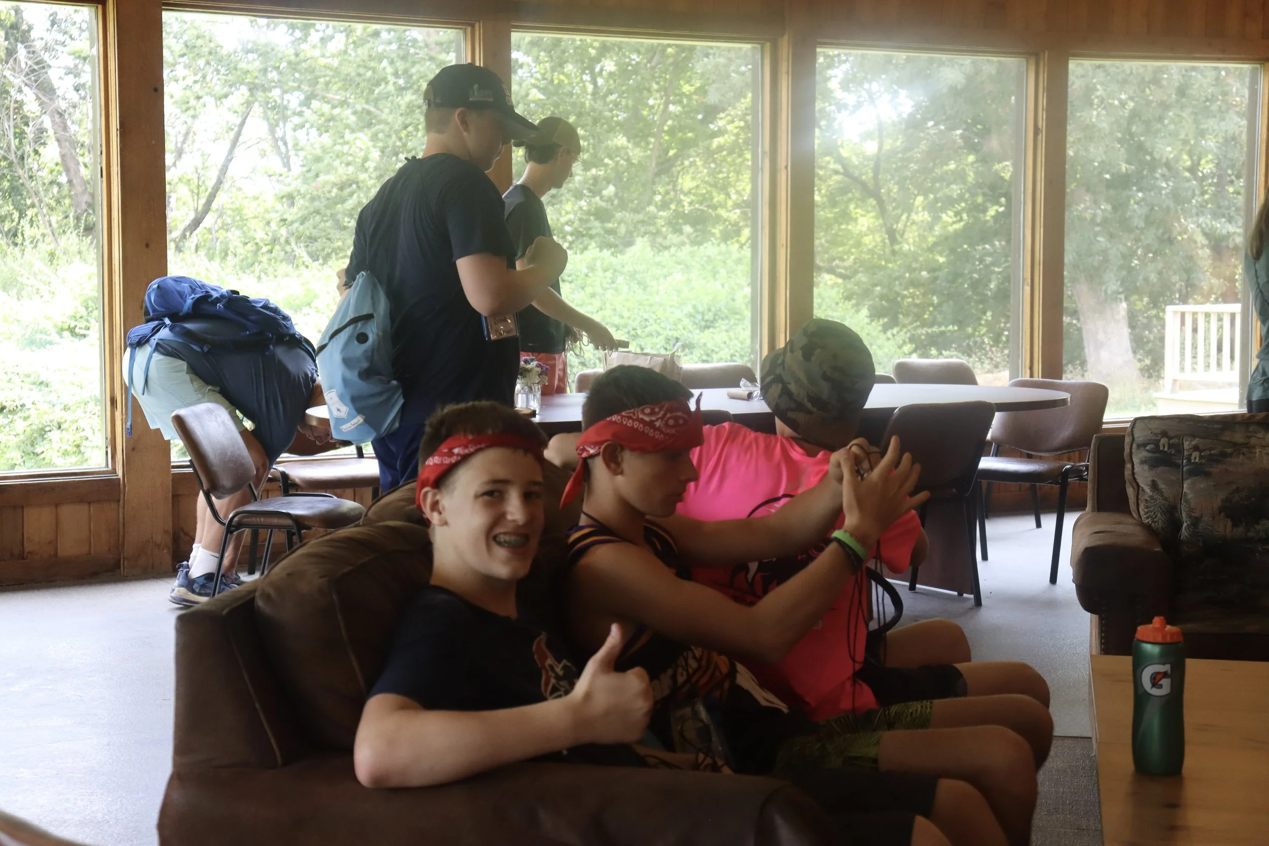 A group of young people sitting and standing inside a room with large windows, with a view of green trees outside. Some of them are wearing red bandanas and some have backpacks.