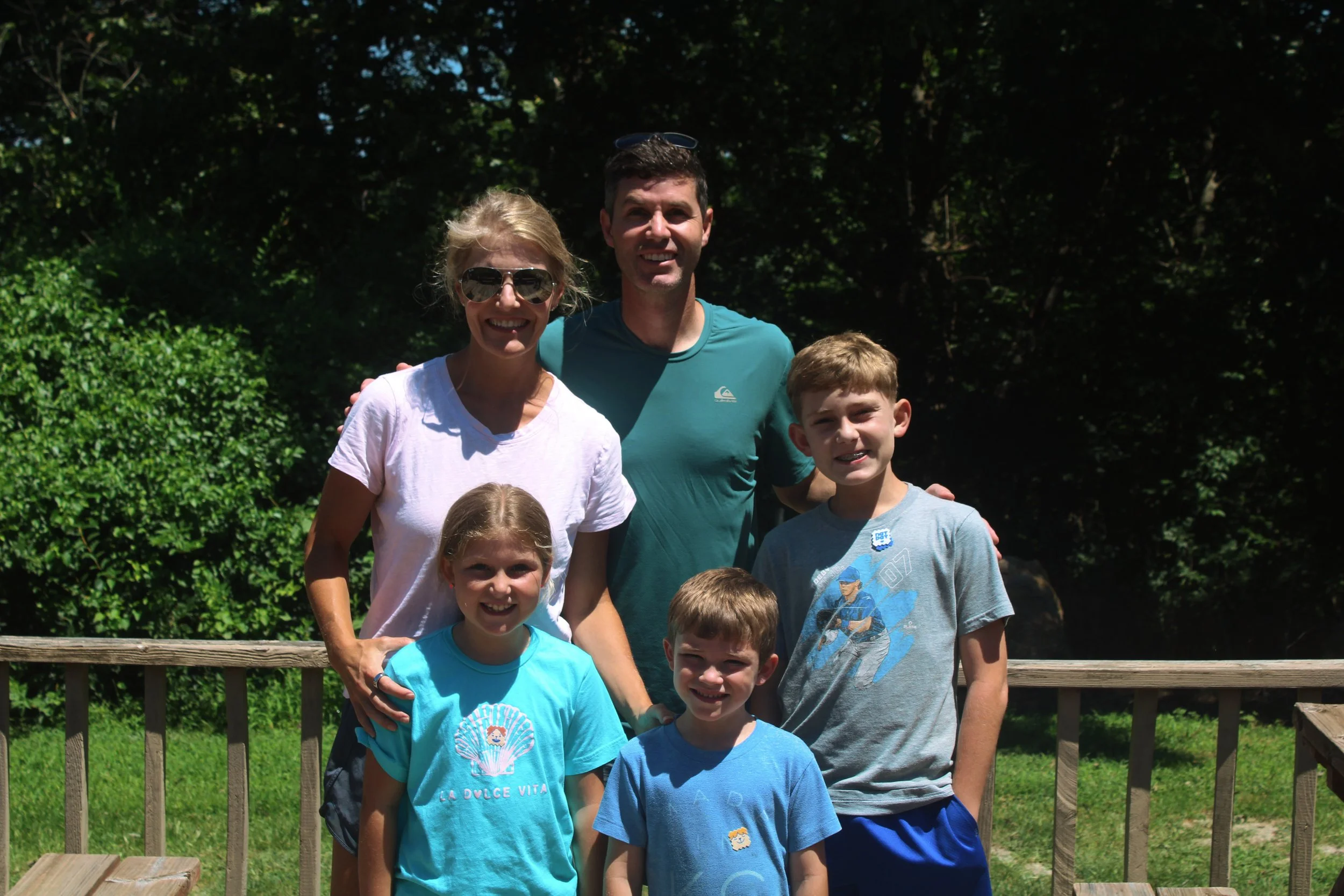 A group of smiling people, including children and adults, standing outdoors behind a wooden fence with lush green trees in the background.
