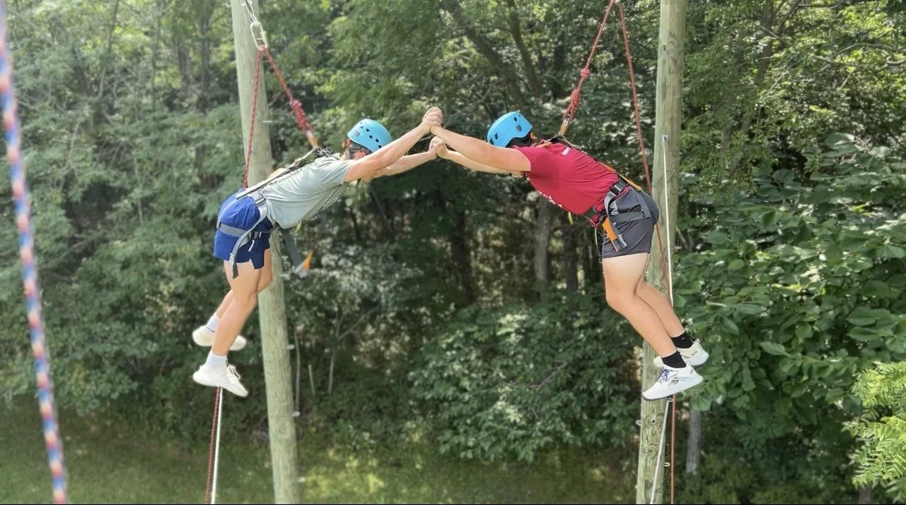 Two children wearing helmets and harnesses are reaching across a gap on an outdoor ropes course, touching hands with each other.