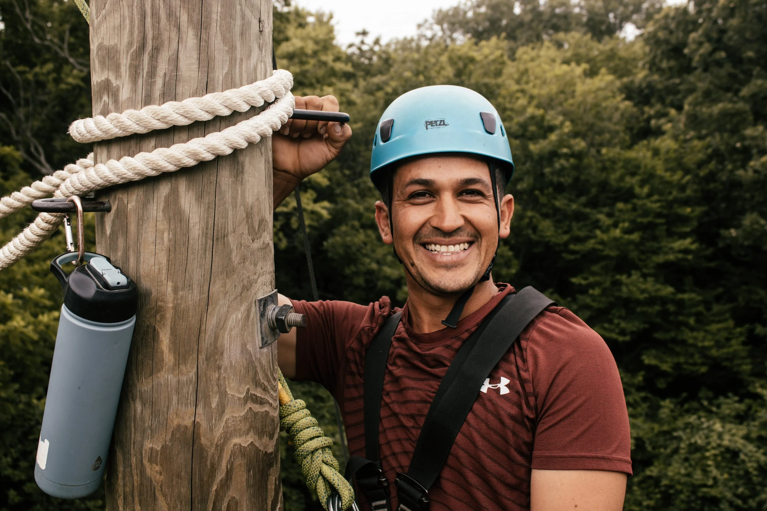 A man wearing a blue safety helmet and a maroon shirt is smiling as he stands next to a wooden utility pole with climbing equipment, including a rope, clips, and a water bottle attached to the pole, surrounded by greenery.