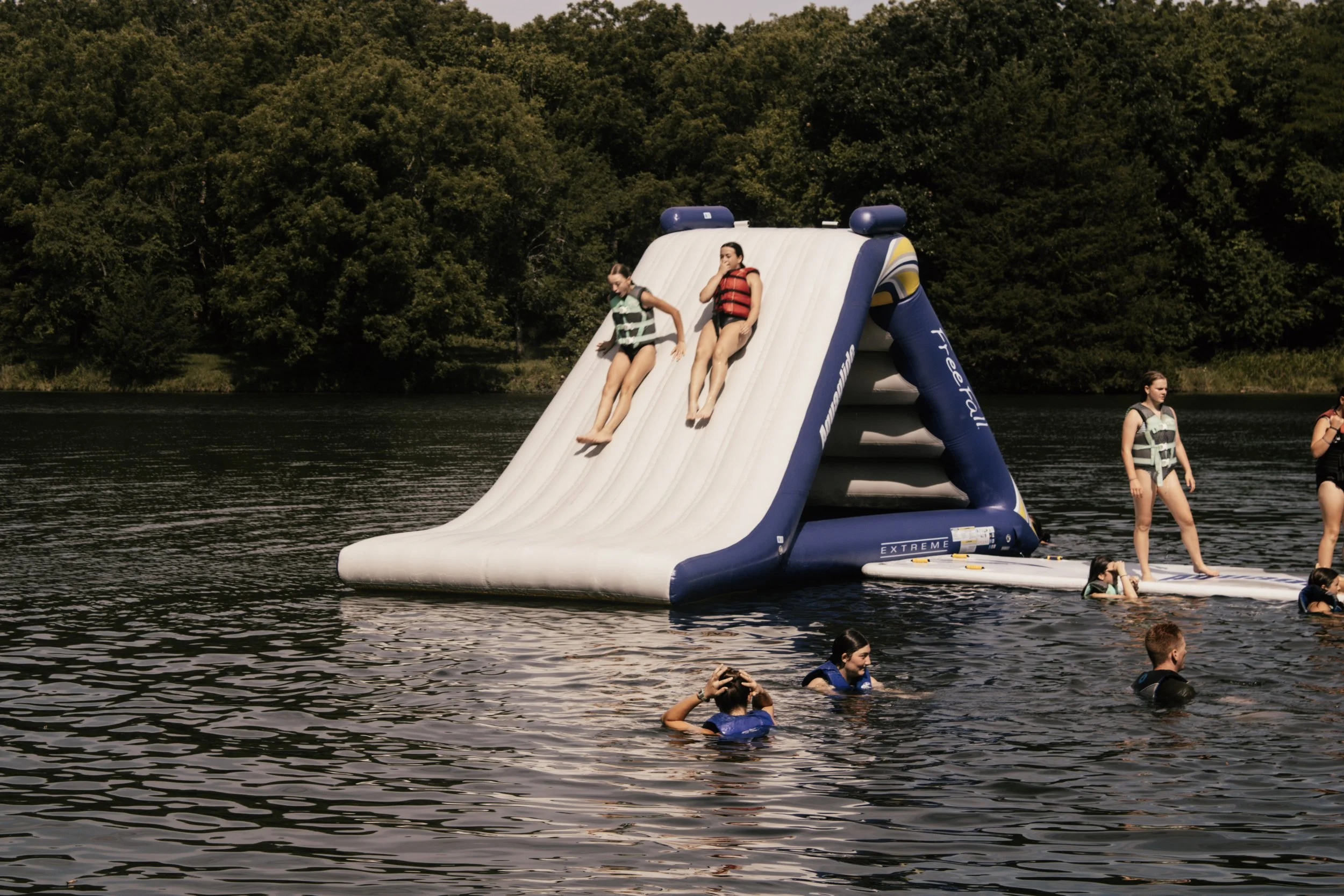 Two children in life jackets slide down an inflatable water slide into a lake while others swim and float nearby, with a background of trees and a clear sky.
