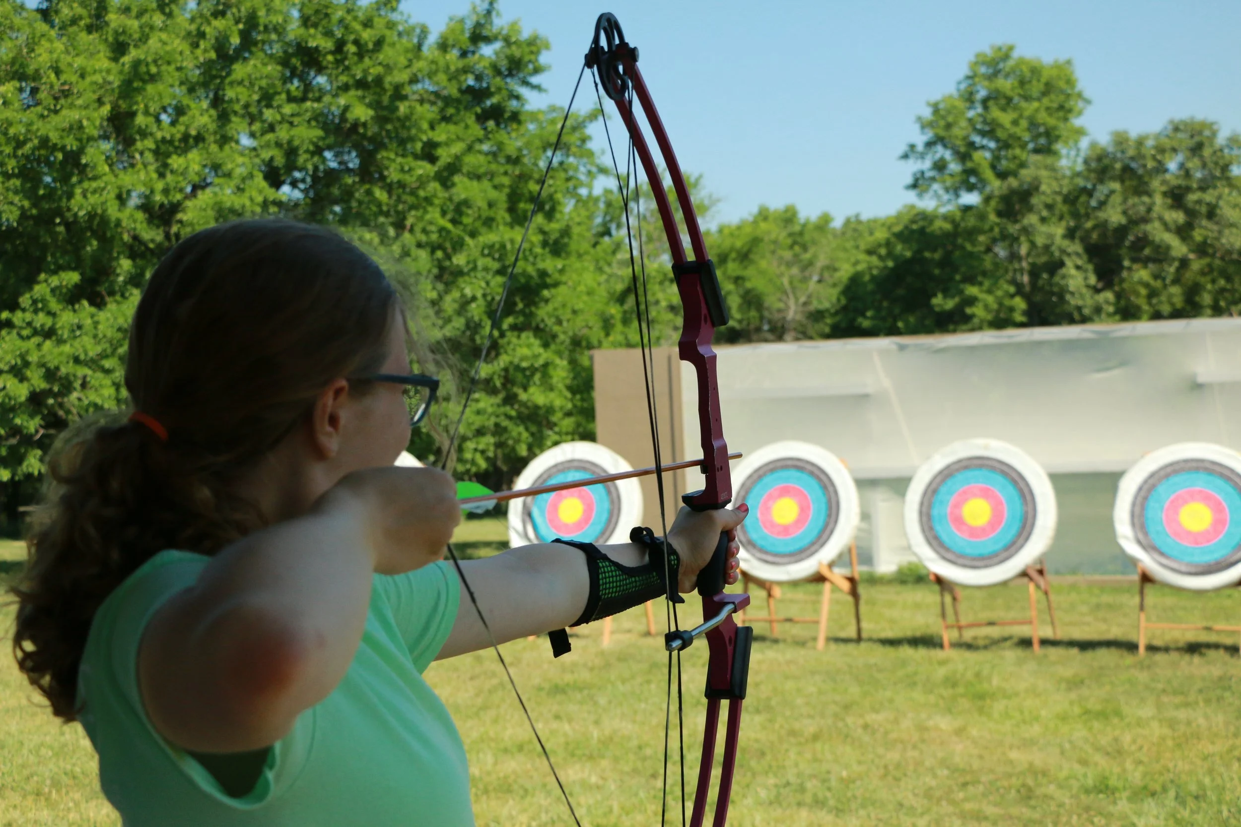 A woman practicing archery outdoors, aiming her bow at target targets with colorful concentric circles in the background, with trees and a blue sky.