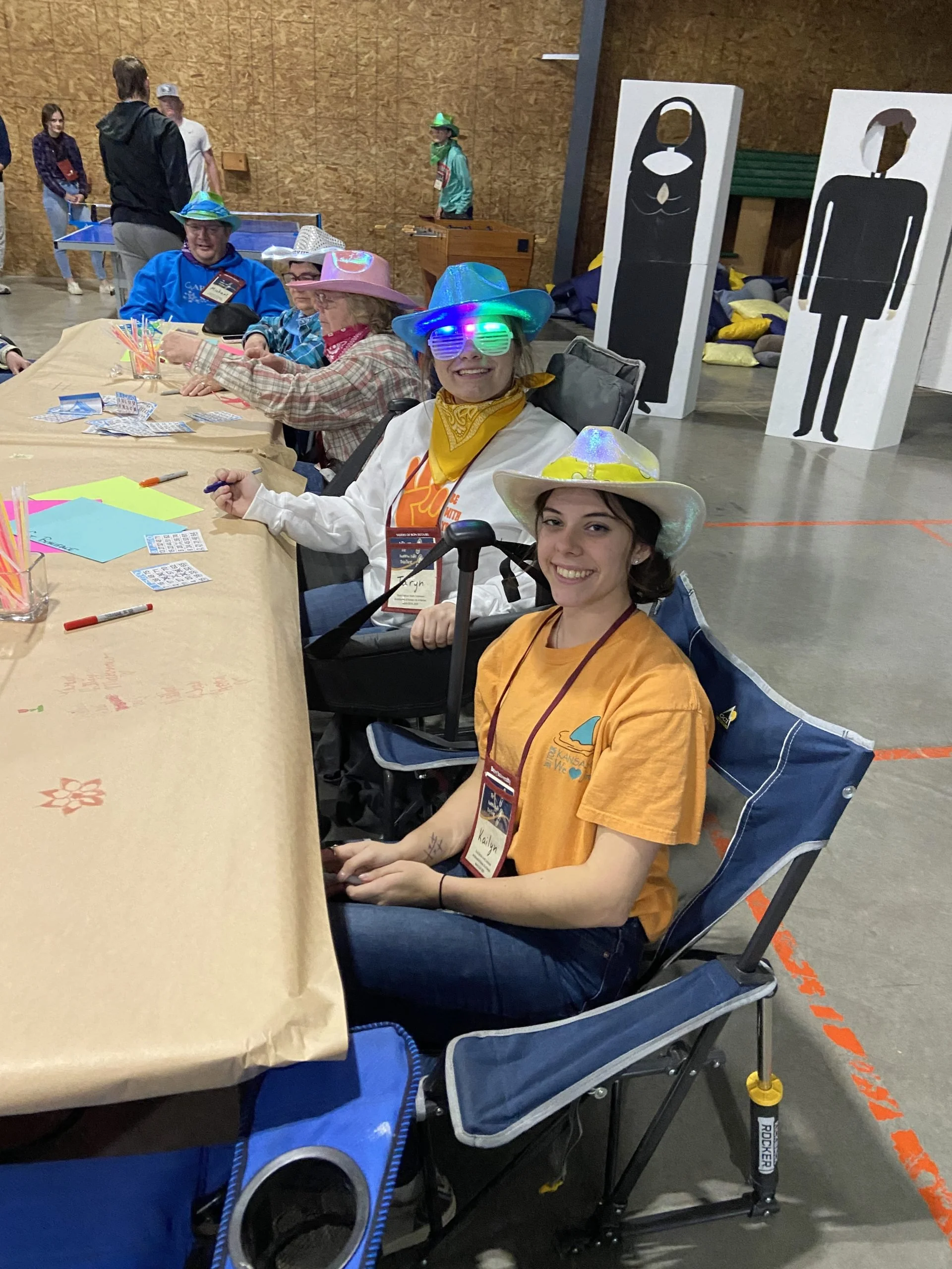 People sitting at a table in a convention or event space, wearing colorful hats and costumes, with signs in the background depicting abstract human figures.
