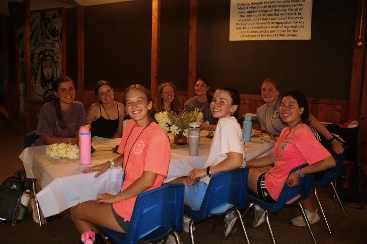 Eight young women sitting around a table in a dining hall, smiling and posing for the photo. The table has flowers and water bottles on it, and everyone appears happy and relaxed.