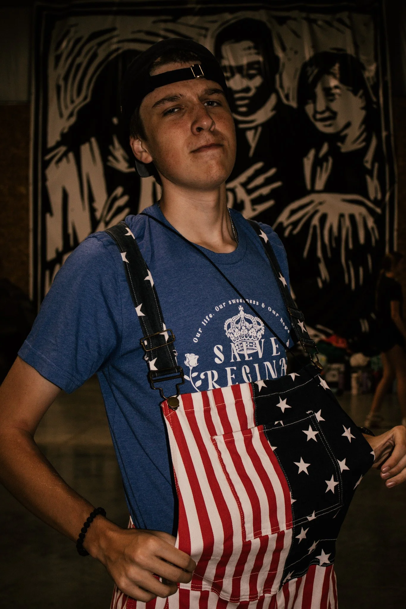 Young man wearing a blue t-shirt, a backward black cap, and an American flag-themed apron, standing indoors in front of a black and white portrait artwork.