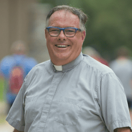 Clergy member smiling outdoors, wearing glasses and a clerical collar