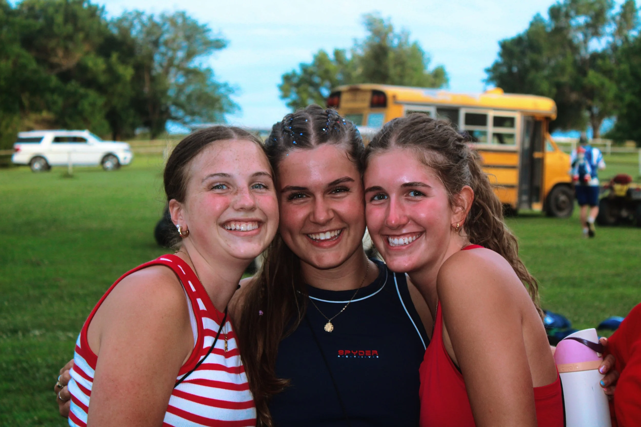 Three young women smiling and hugging outdoors, with a yellow school bus, a white vehicle, and trees in the background.