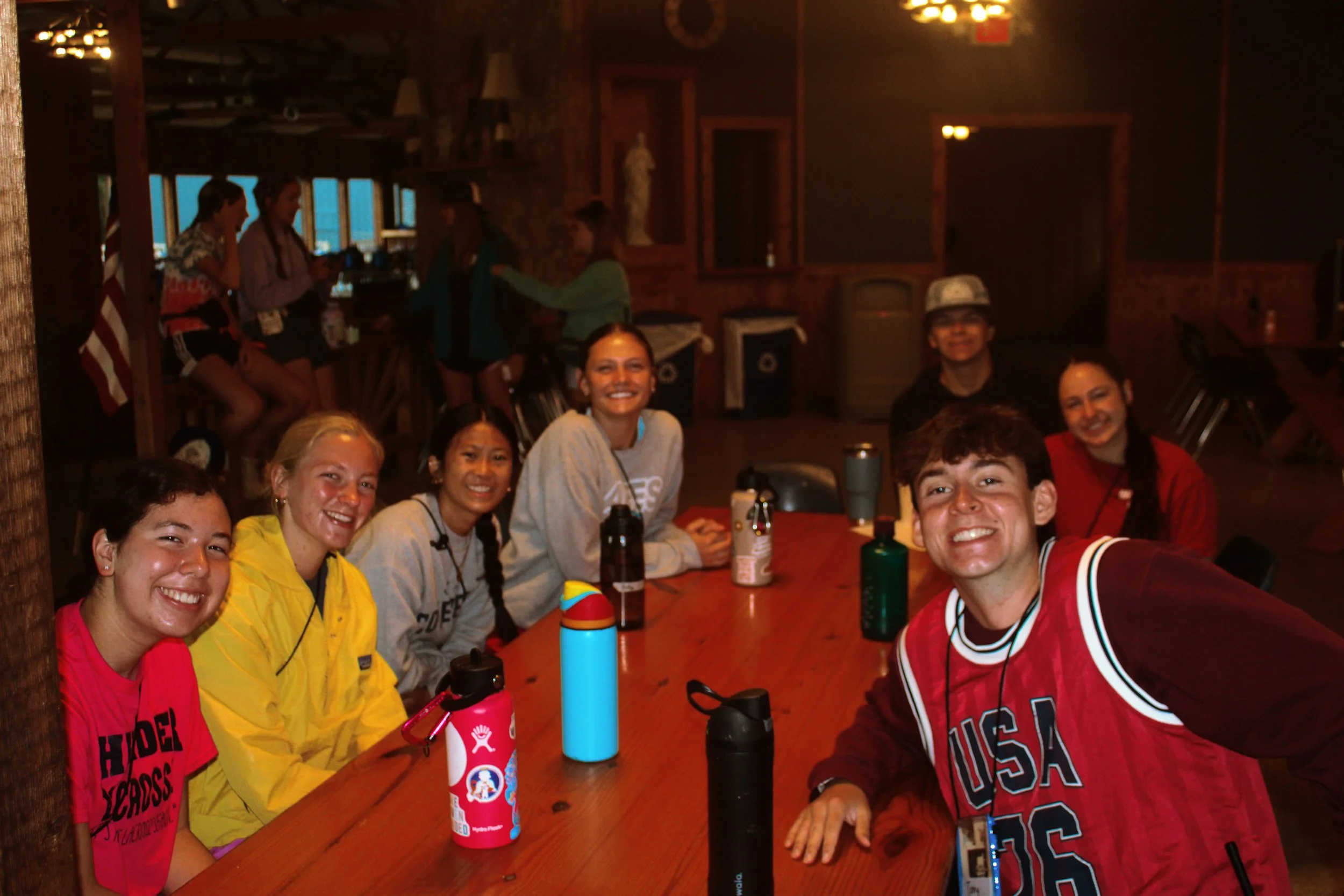 A group of smiling young adults sitting around a wooden table in a dimly lit rustic restaurant or bar. Some are wearing colorful casual clothing including yellow, pink, gray, red, and black. There are water bottles, a cup with straws, and personal it
