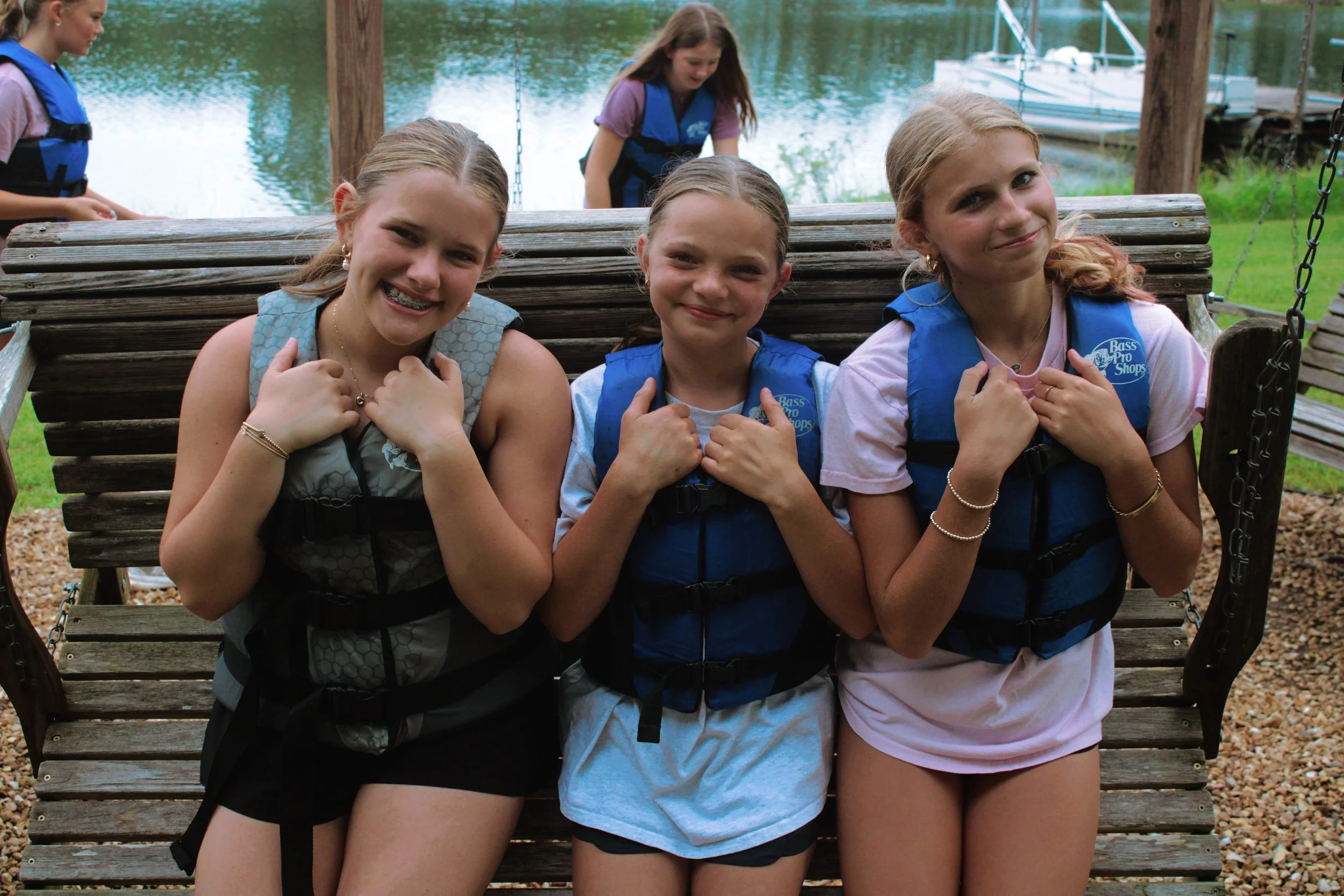 Three girls sitting on a wooden park bench near a lake, smiling and giving thumbs-up signs, with two girls in the background near the water.