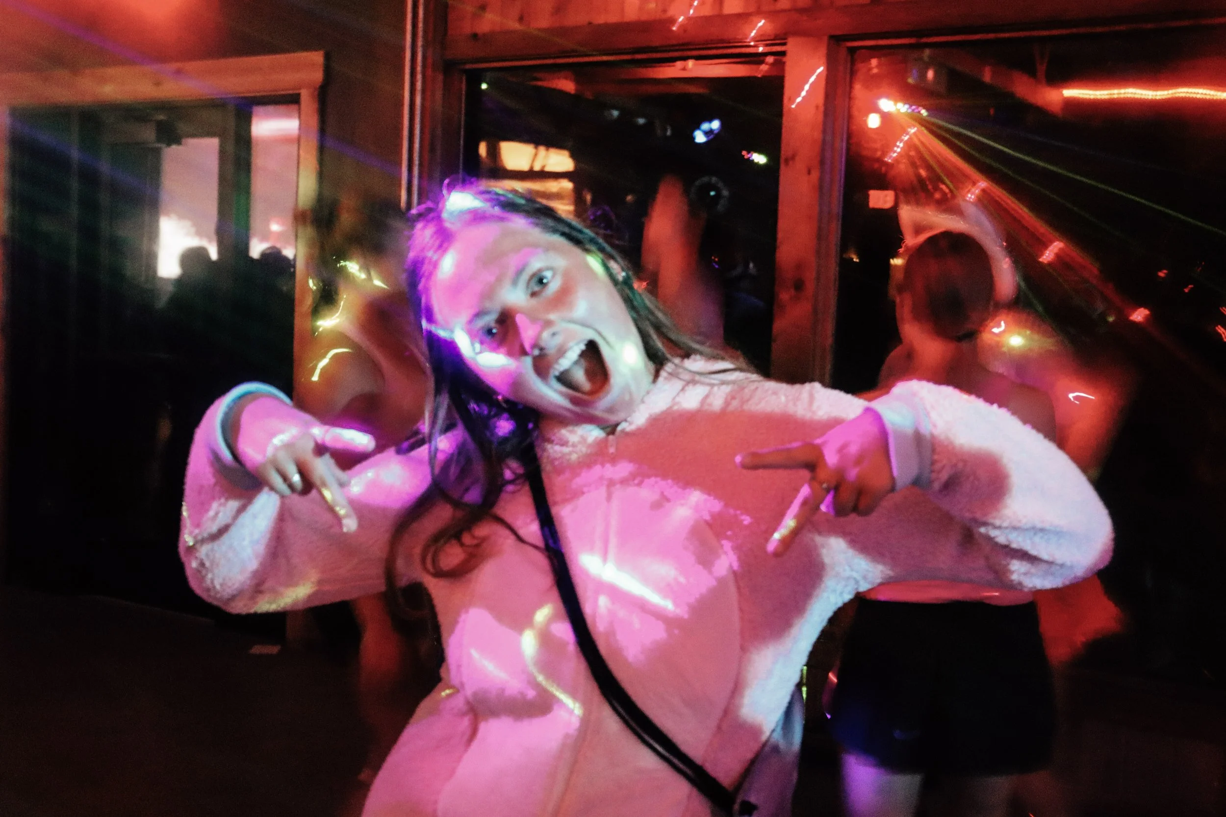 A woman with shoulder-length hair, wearing a fuzzy pink sweater, is excitedly posing for a photo with her mouth open and pointing with her fingers. There are colorful lights projecting patterns on her face and sweater, and a person with short hair an