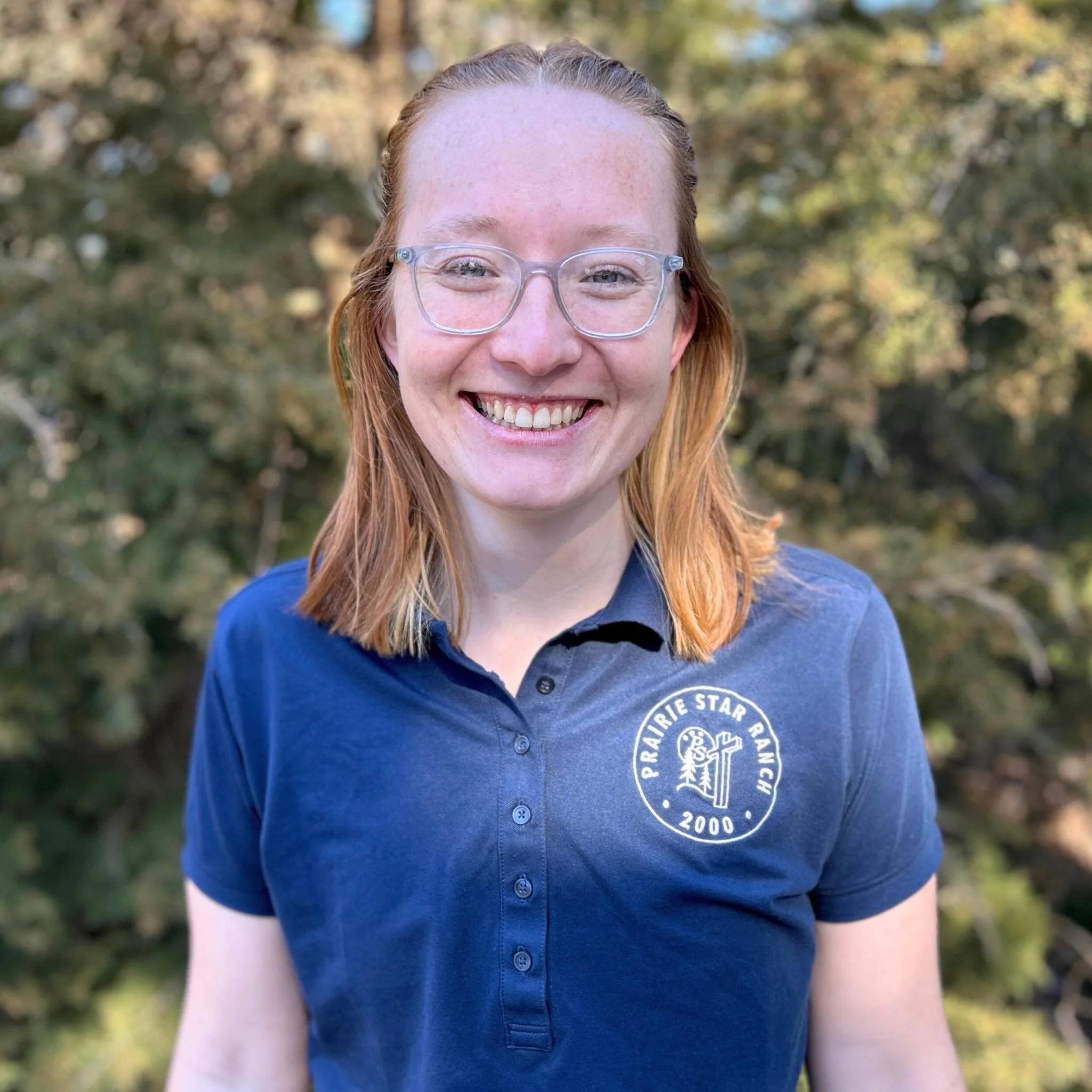 Close-up of a woman with red hair and glasses smiling outdoors, wearing a blue shirt with a circular logo.