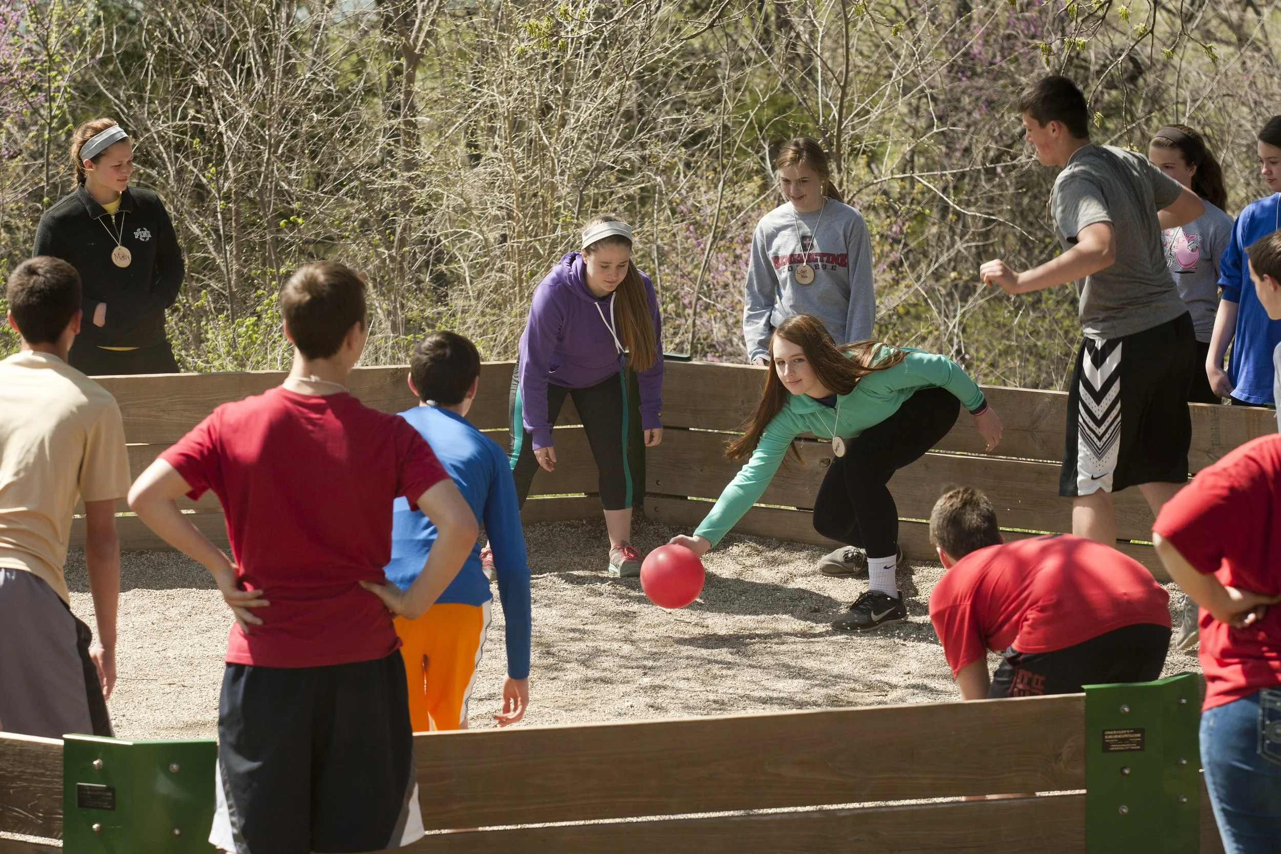 Group of children outdoors playing a game with a red ball, some children are standing inside a wooden enclosure and others are watching nearby, trees and pink flowers in the background.