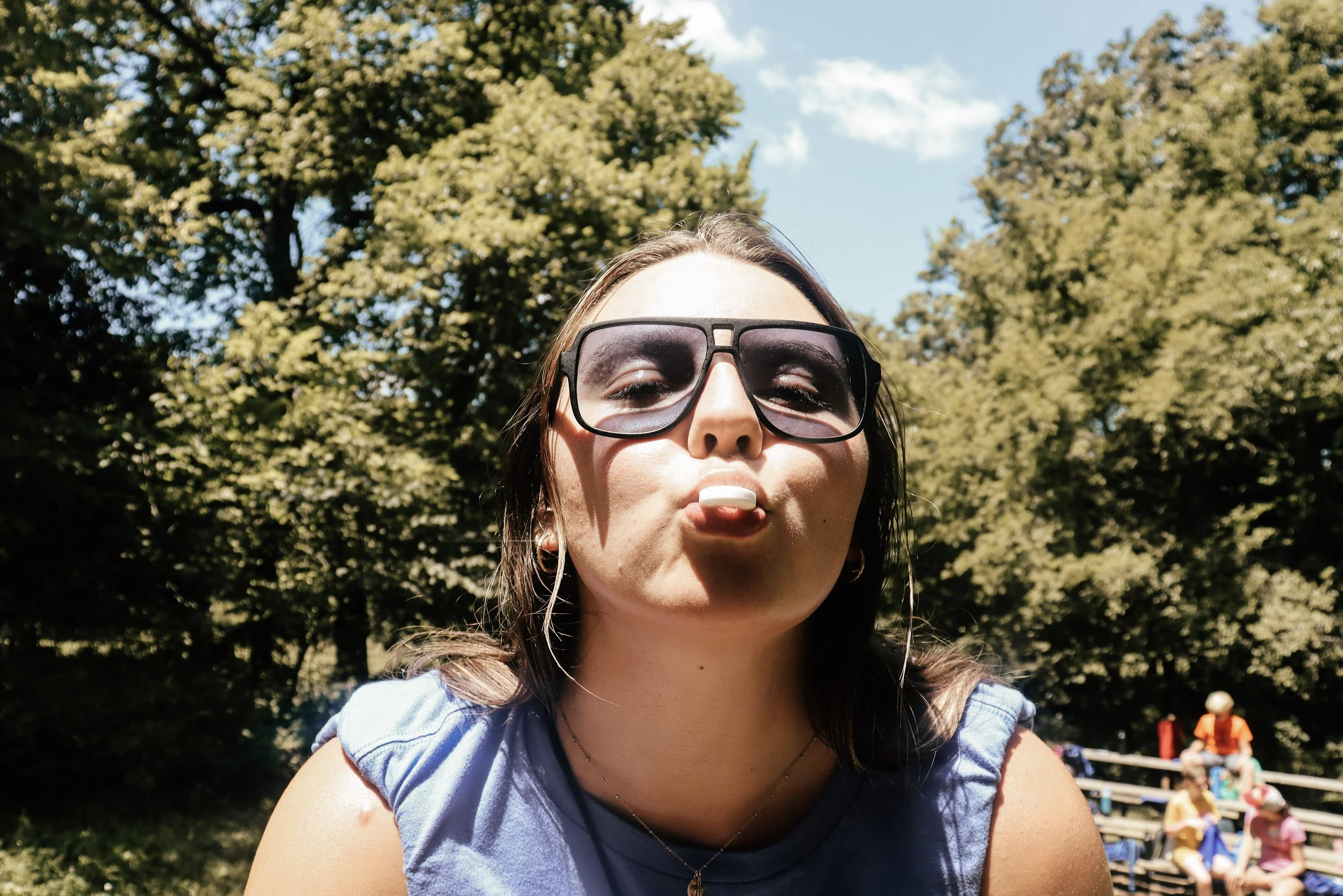 Close-up of a woman wearing sunglasses and blowing a bubble with bubble gum outdoors on a sunny day, with trees and people in the background.