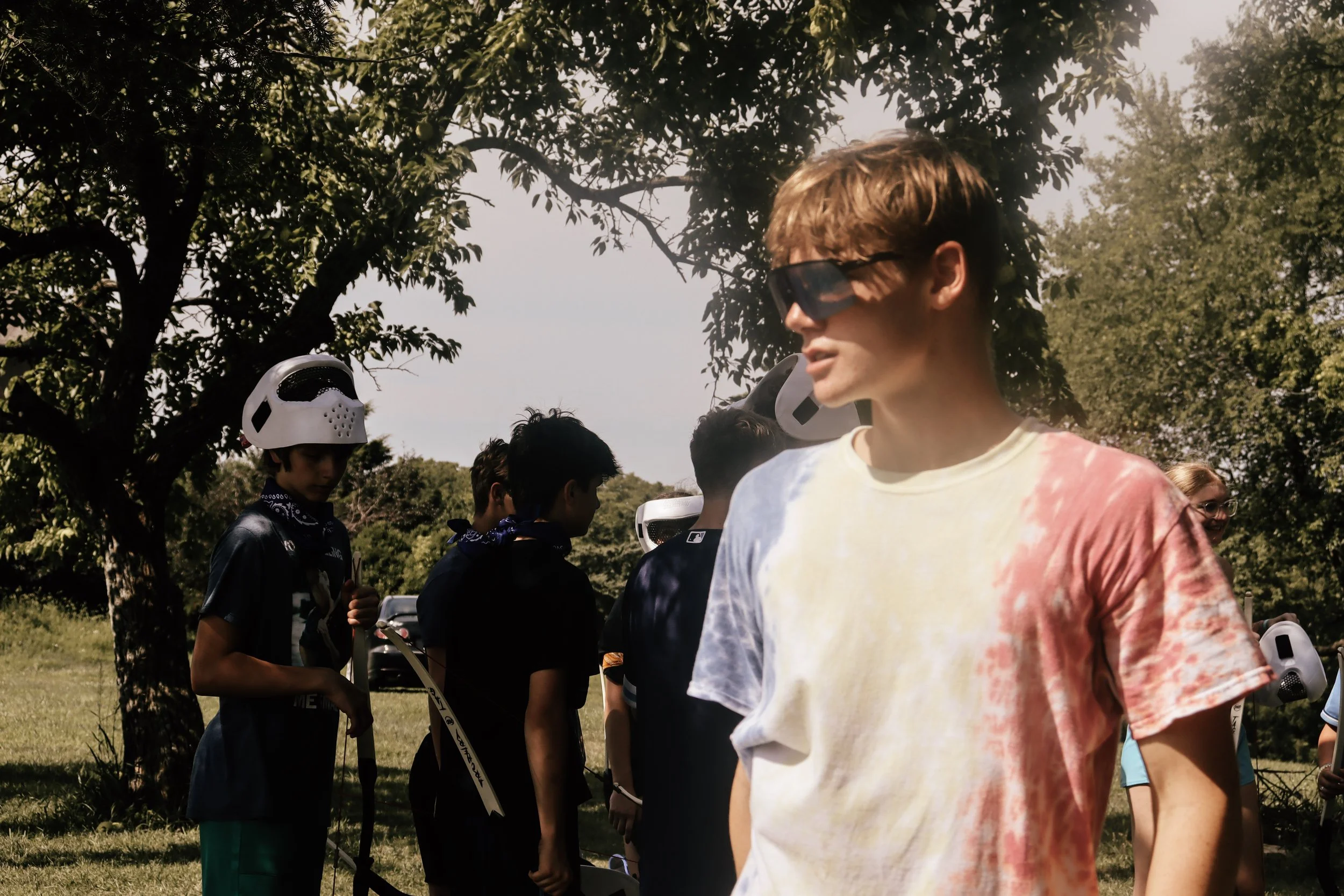 Group of children and teenagers outdoors, some wearing helmets and sunglasses, gathered under trees on a sunny day.