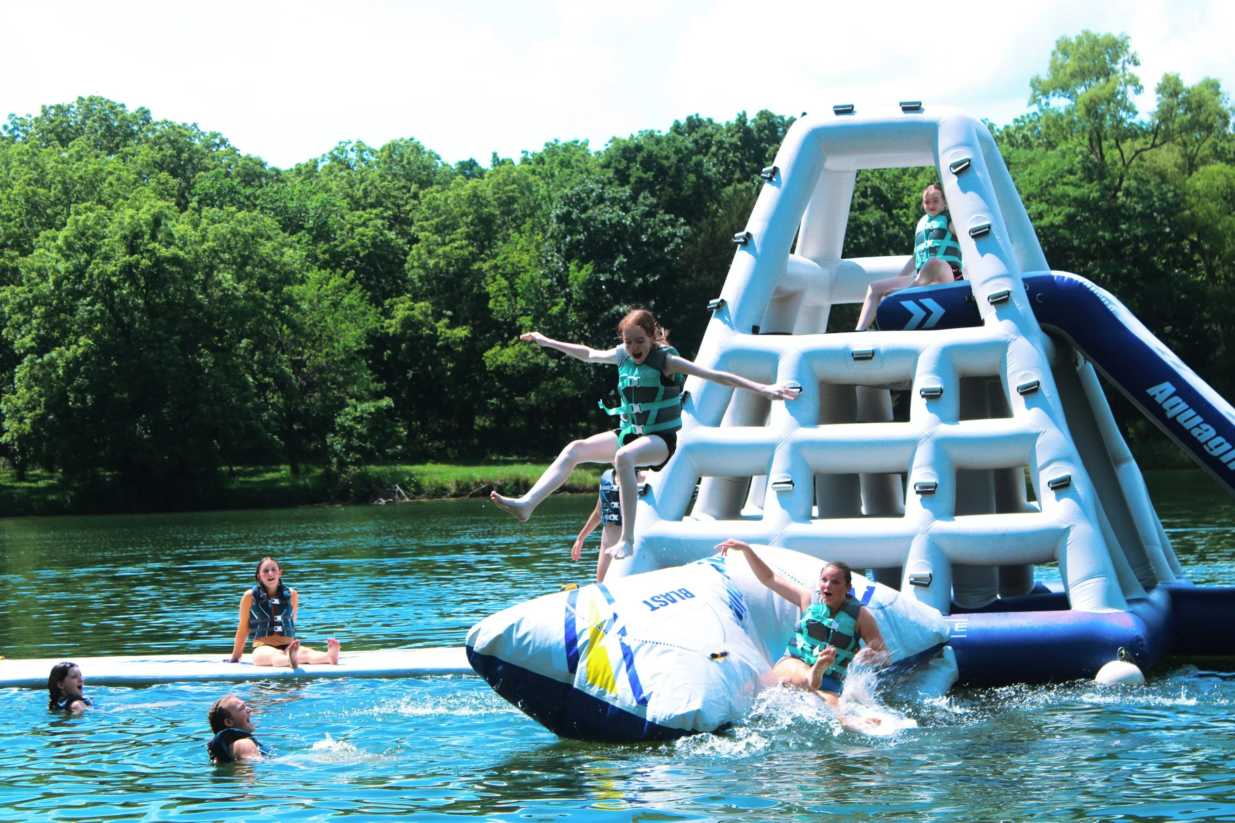 Children enjoying a summer day on an inflatable water slide set up on a lake, with some climbing up the slide and others jumping and playing in the water.